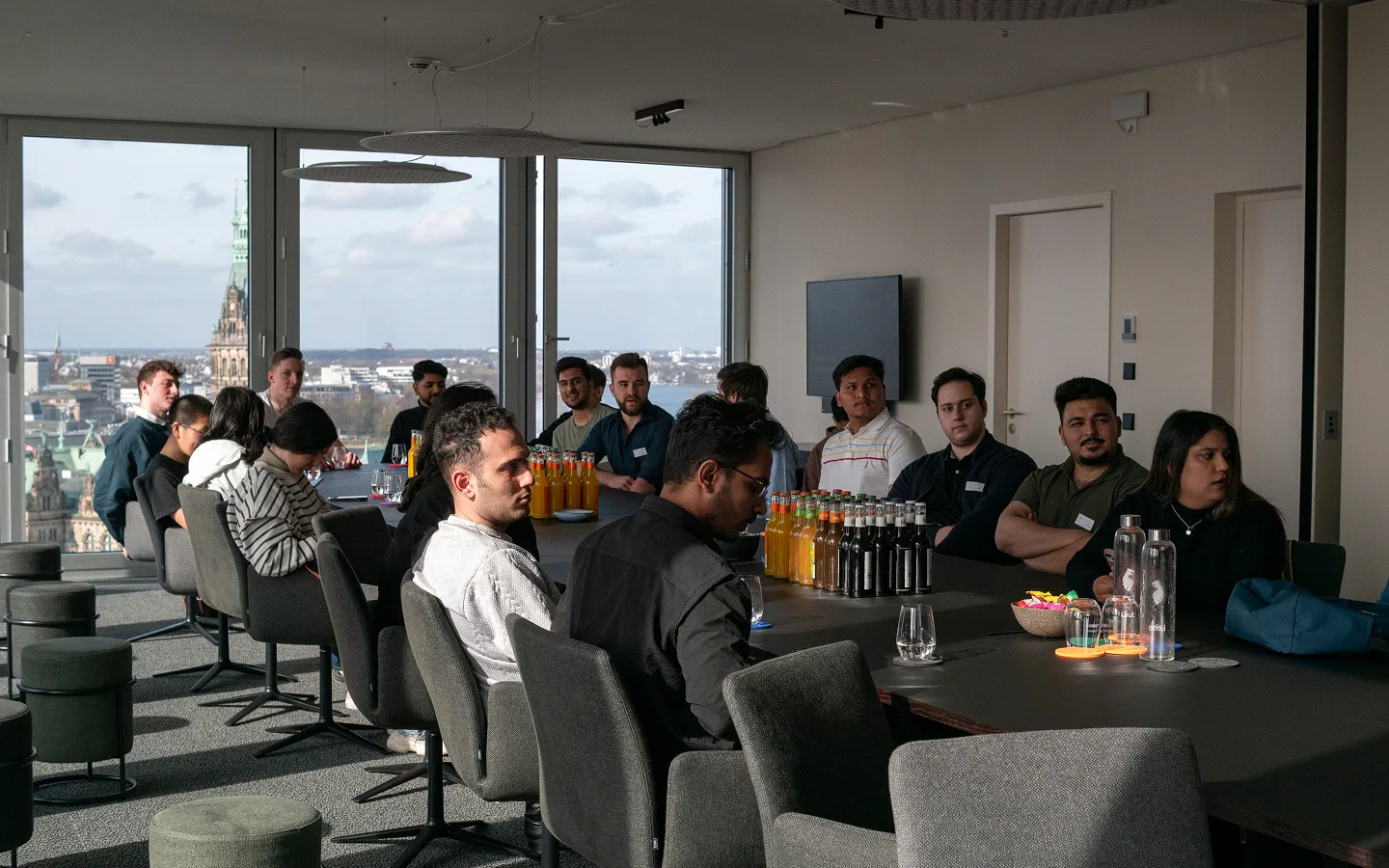 Group of people seated around a conference table with drinks, in a room with large windows showing a cityscape.