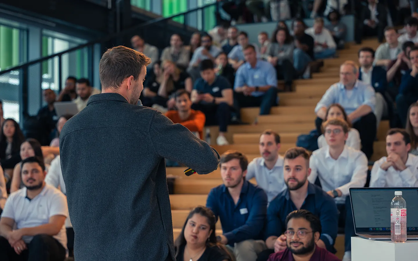 Man speaking to an attentive audience seated on wooden bleachers in a modern indoor setting.
