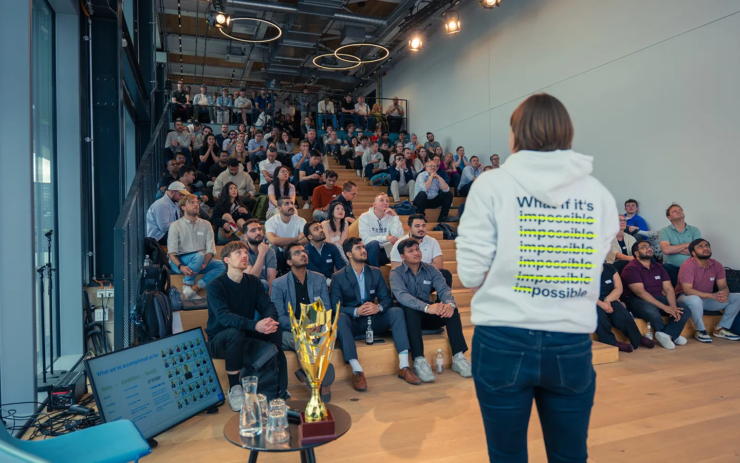 Speaker in a white hoodie with text saying 'What if it's impossible' repeated and crossed out, presenting to a large audience seated on tiered wooden steps in a modern room.