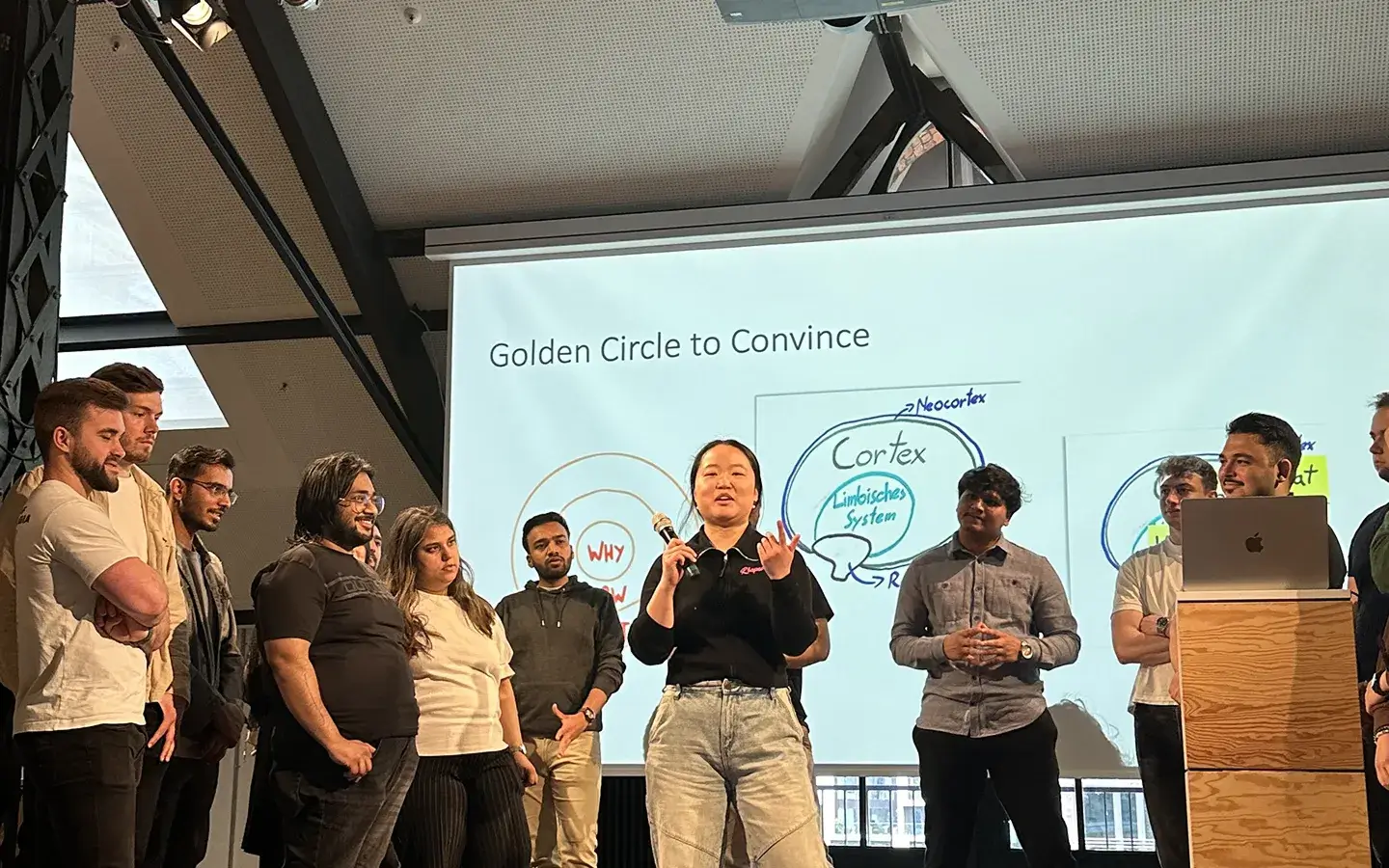 A group of people standing and listening to a presenter holding a microphone in front of a presentation slide titled 'Golden Circle to Convince'.