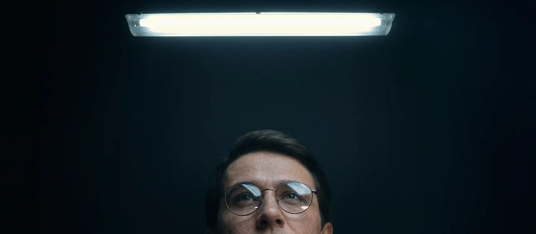 Close-up of a man wearing round glasses looking up at a bright fluorescent light against a dark background.