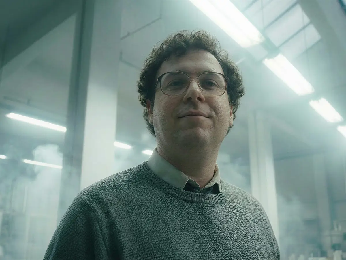 Man with curly hair and glasses wearing a gray sweater in a foggy industrial setting with fluorescent lights.