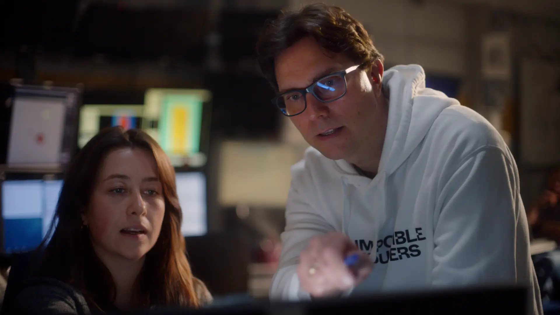 A man in glasses and a white hoodie pointing at a screen while a woman with long brown hair watches attentively, with computer monitors in the background.