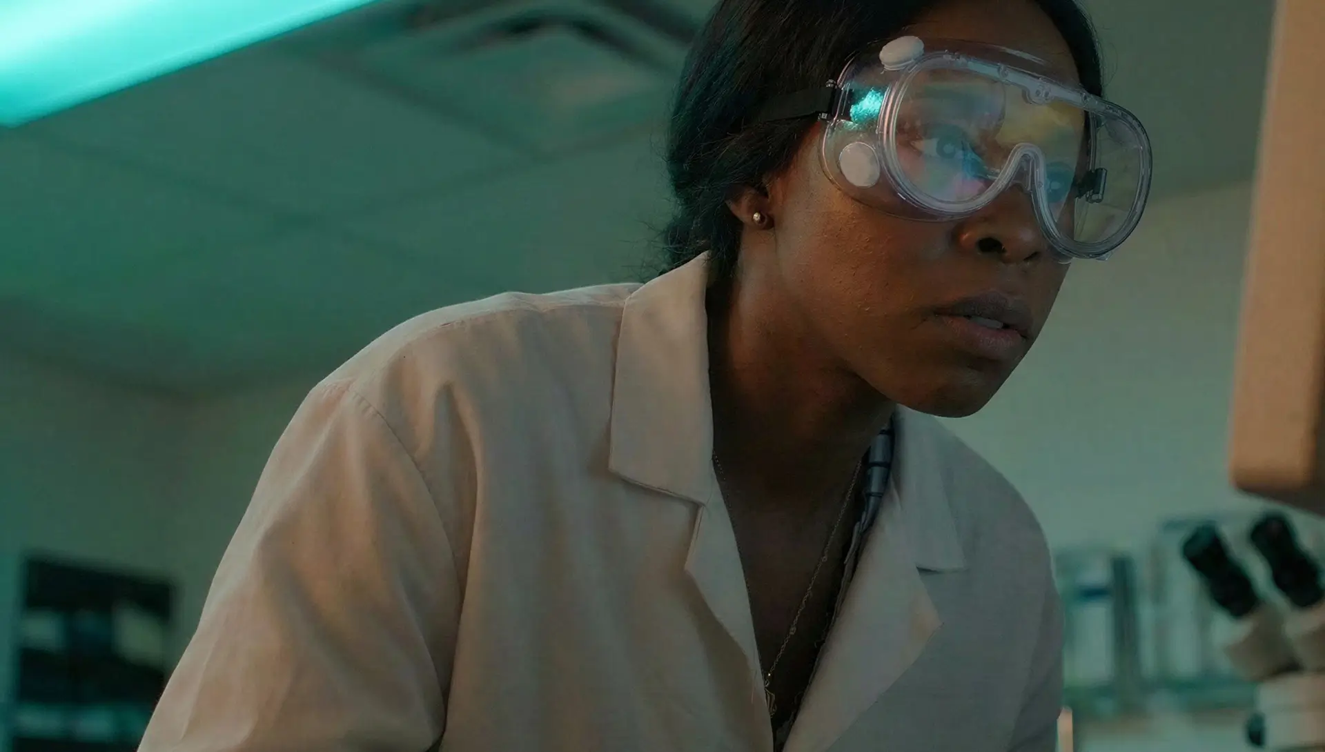 Woman in a lab coat and safety goggles looking intently at a microscope in a laboratory.