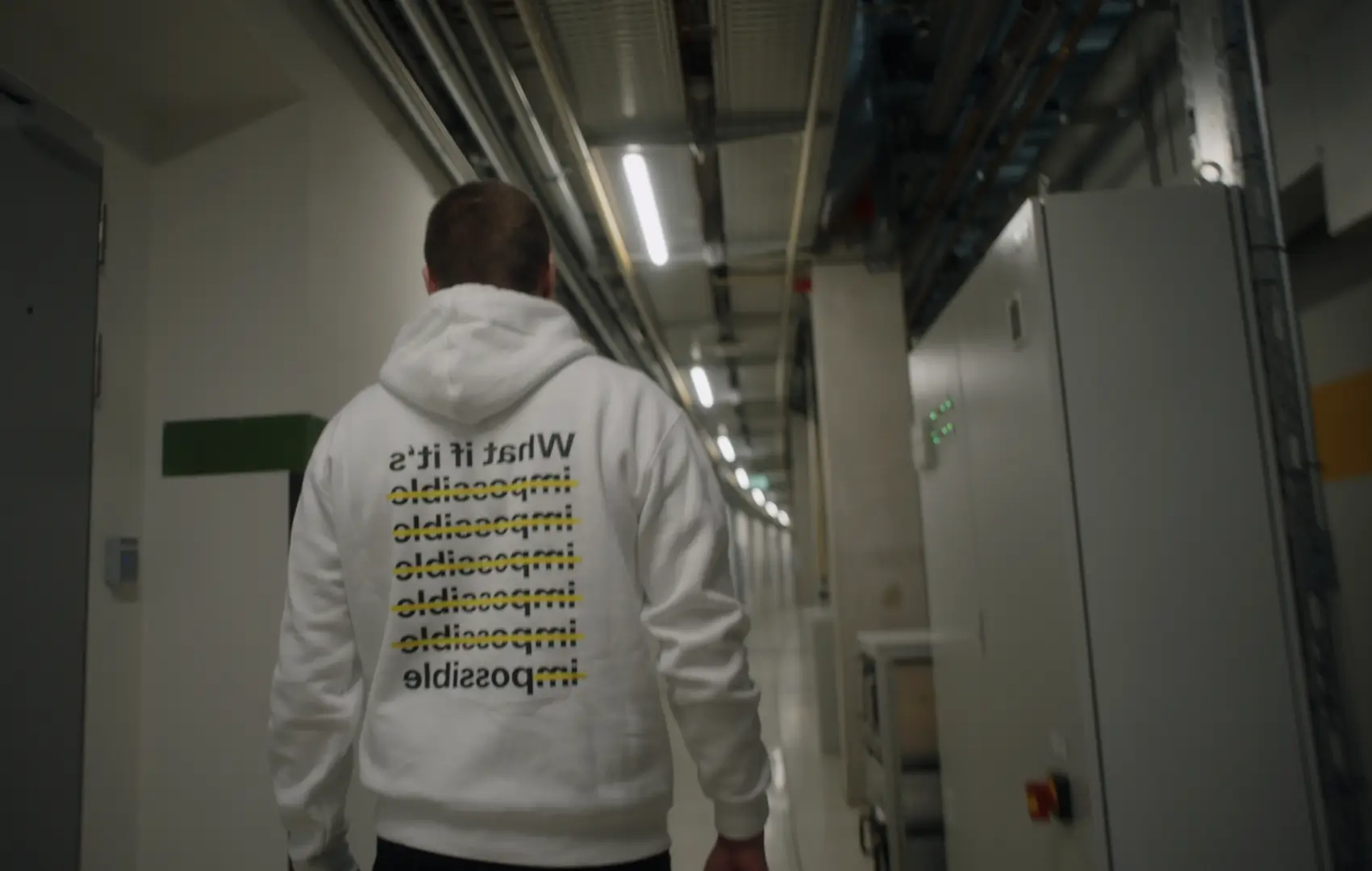 Person walking down a corridor wearing a white hoodie with text 'What if it’s impossible' repeated and crossed out multiple times except the last word 'impossible'.