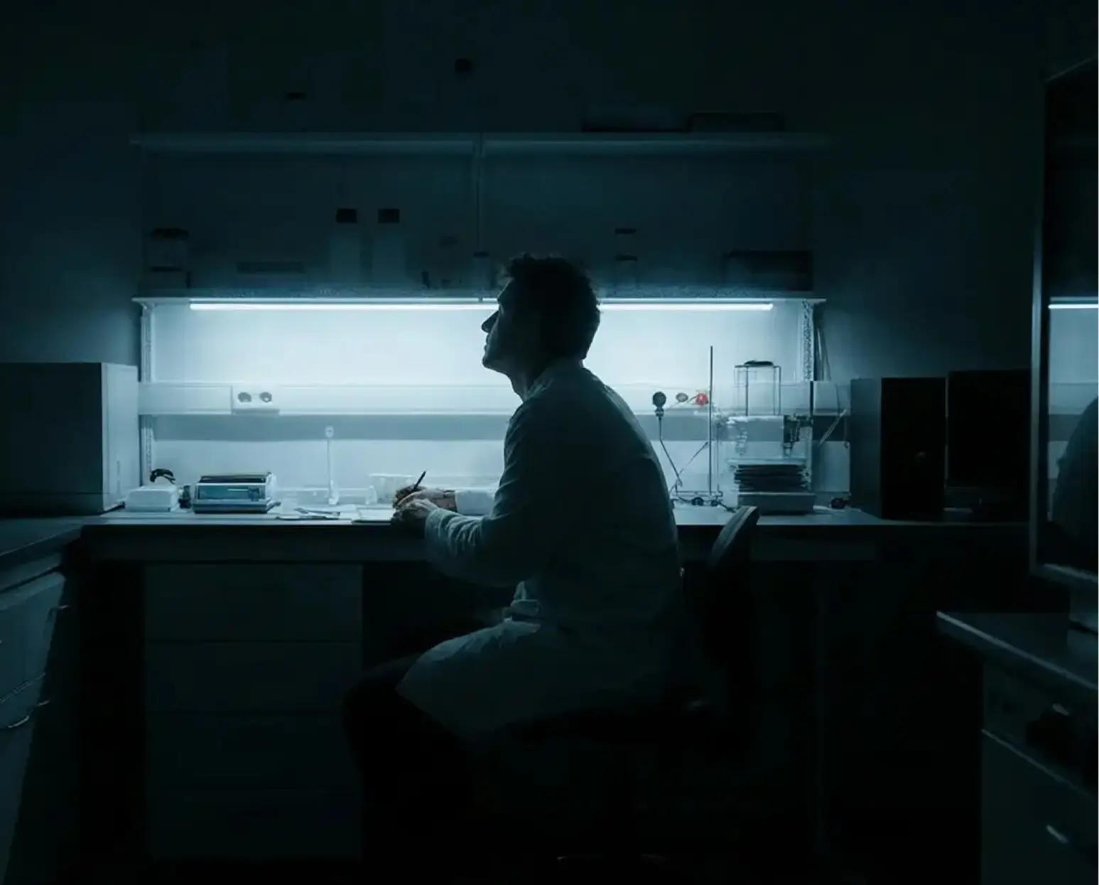 Silhouette of a person in a lab coat sitting at a illuminated laboratory bench writing or drawing.
