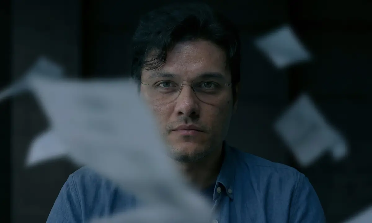 Man with glasses and blue shirt looking directly ahead with blurred flying papers around him against a dark background.