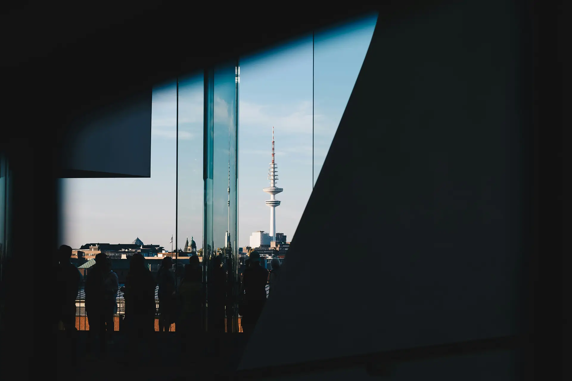 Silhouettes of people inside a building framing a city view with a tall communications tower under a blue sky.