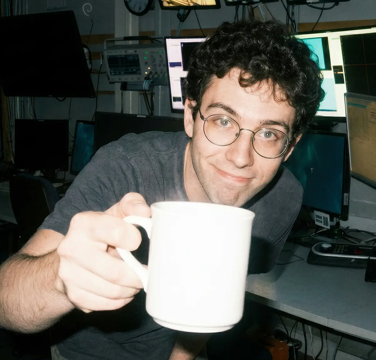 Man with curly dark hair and glasses holding a white mug towards the camera, smiling, with multiple computer monitors in the background.
