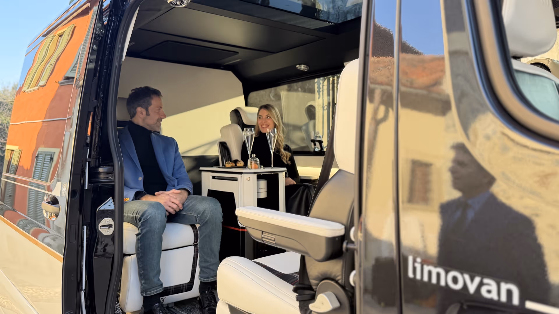 Man and woman seated inside a Moveolux luxury van with drinks on a small table between them - Limovan interior.