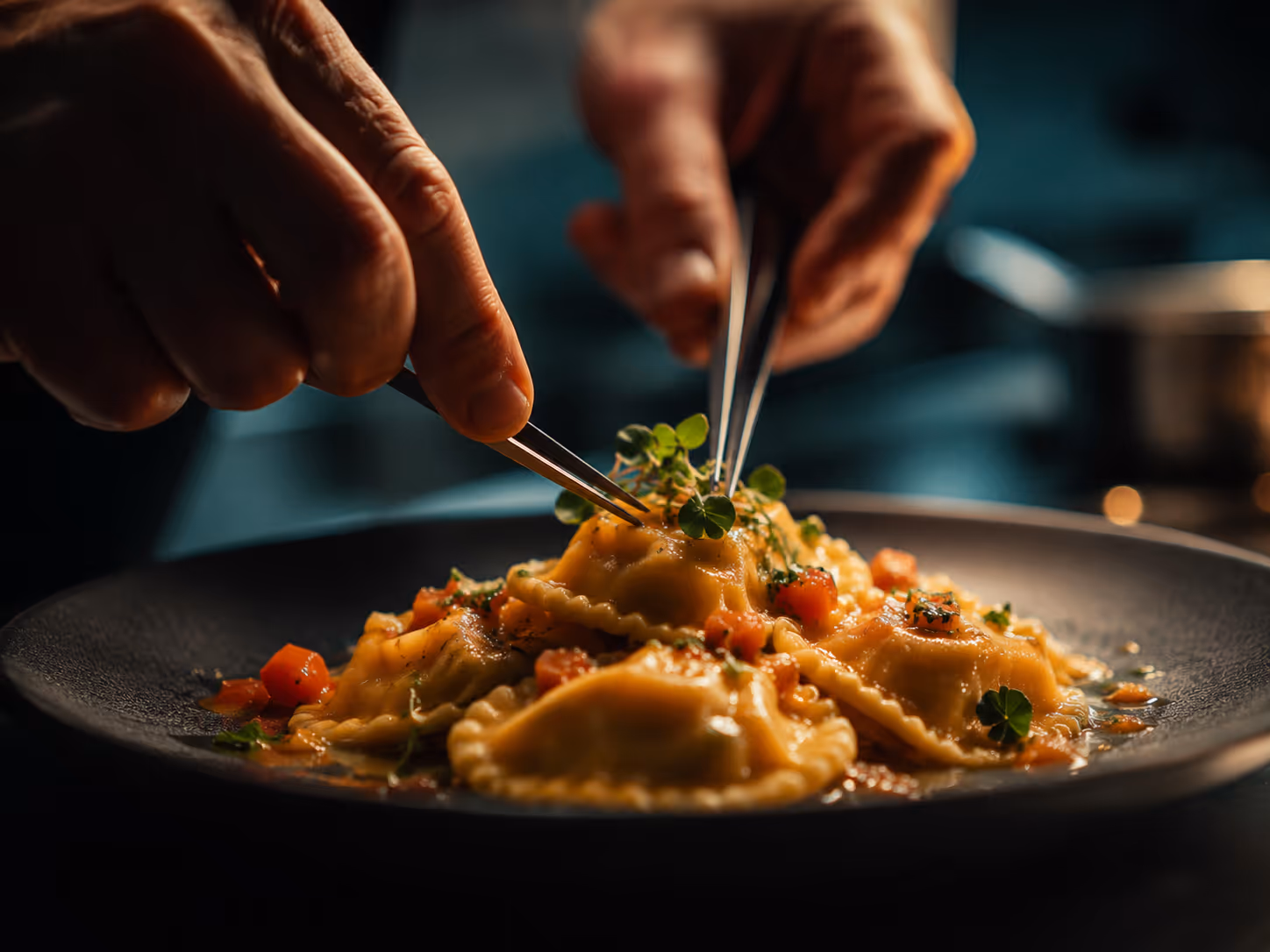 Hands using tweezers to garnish a plate of ravioli pasta with diced tomatoes and herbs.