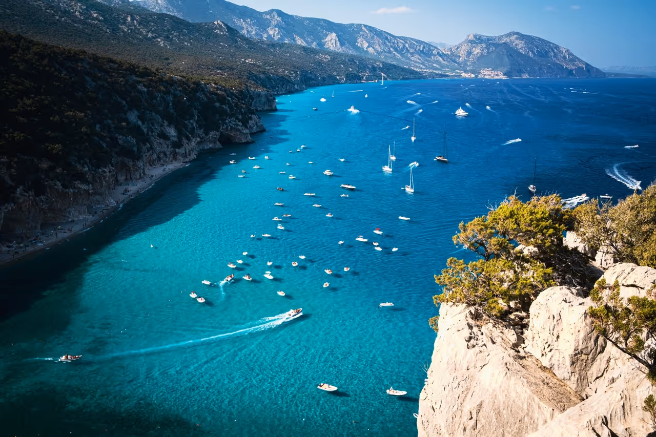 Boats and yachts sailing on clear blue sea near rocky cliffs in Sardinia, a destination for Moveolux yacht charter.