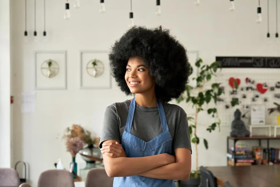 Young woman with an afro hairstyle wearing an apron standing with arms crossed and smiling in a cozy, modern cafe.