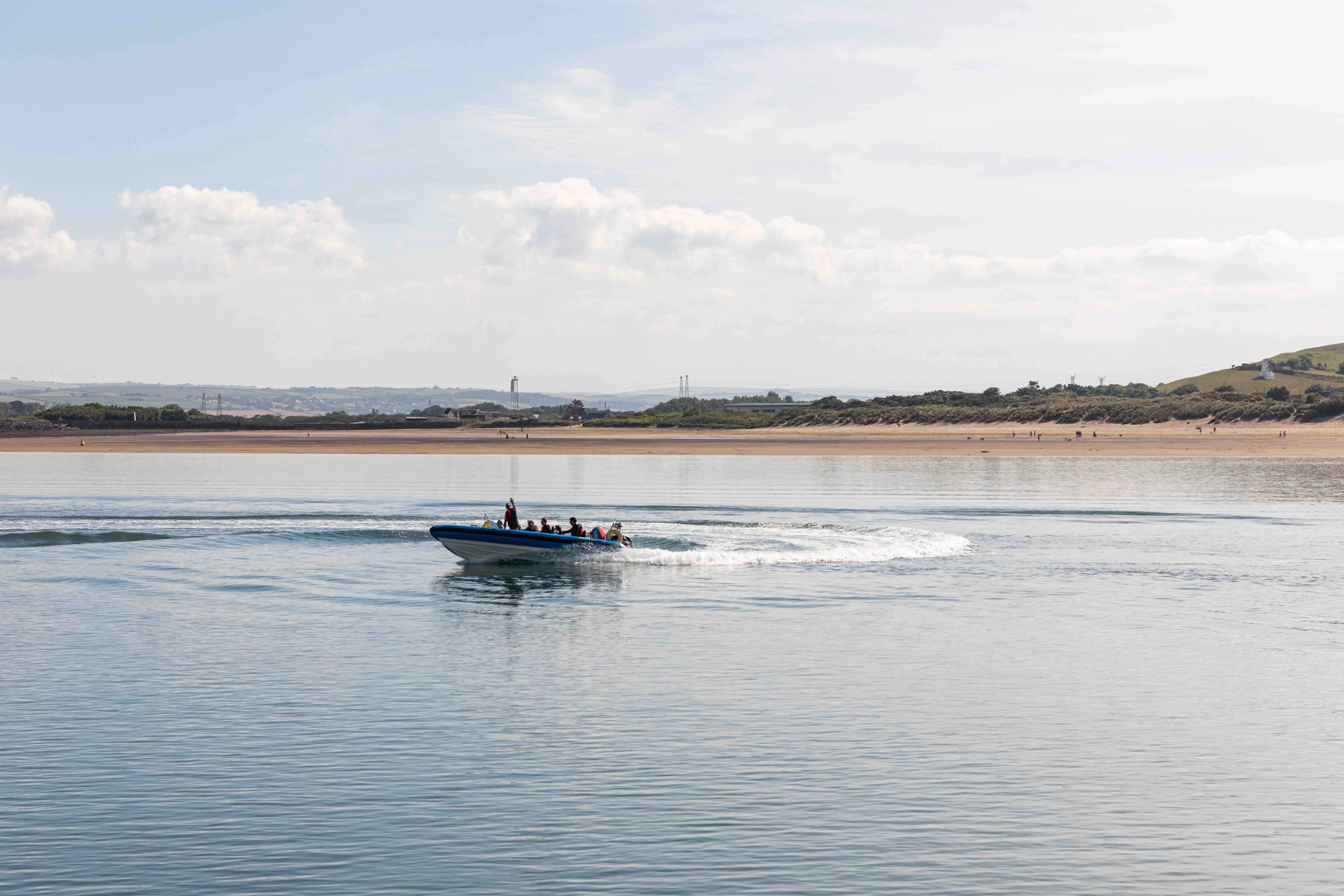 Small boat with several people creating a wake on calm water near a sandy shore with hills and scattered bushes.