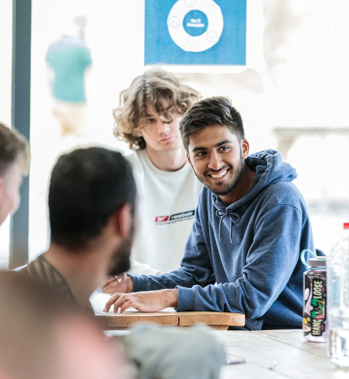 A young man in a blue hoodie smiling as he sits at a table with peers in a bright room.