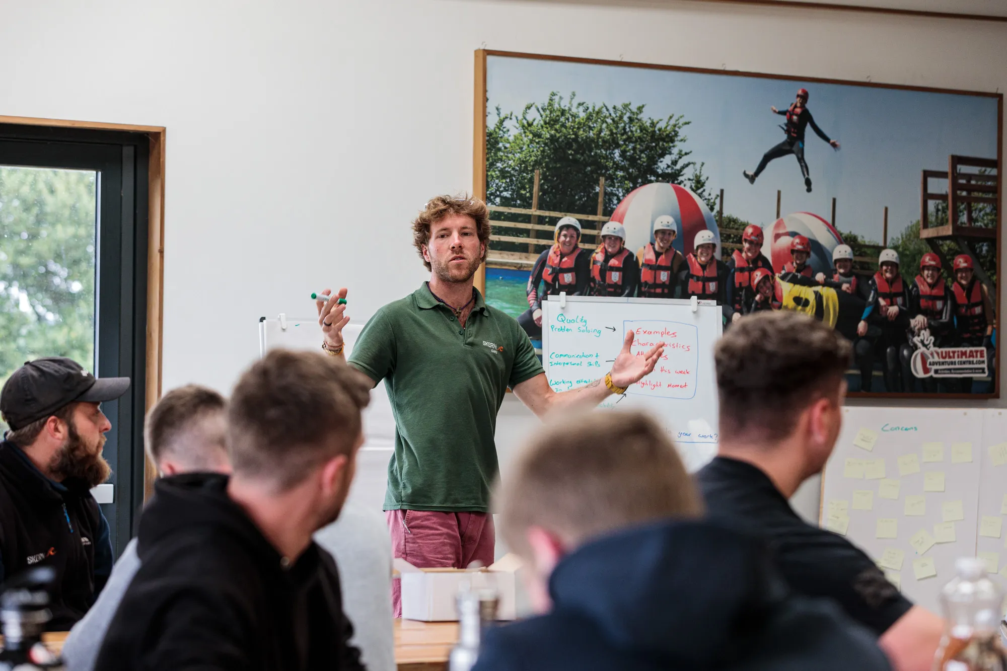 Man in a green polo shirt leading a discussion with a group of people seated around a table in a classroom setting.