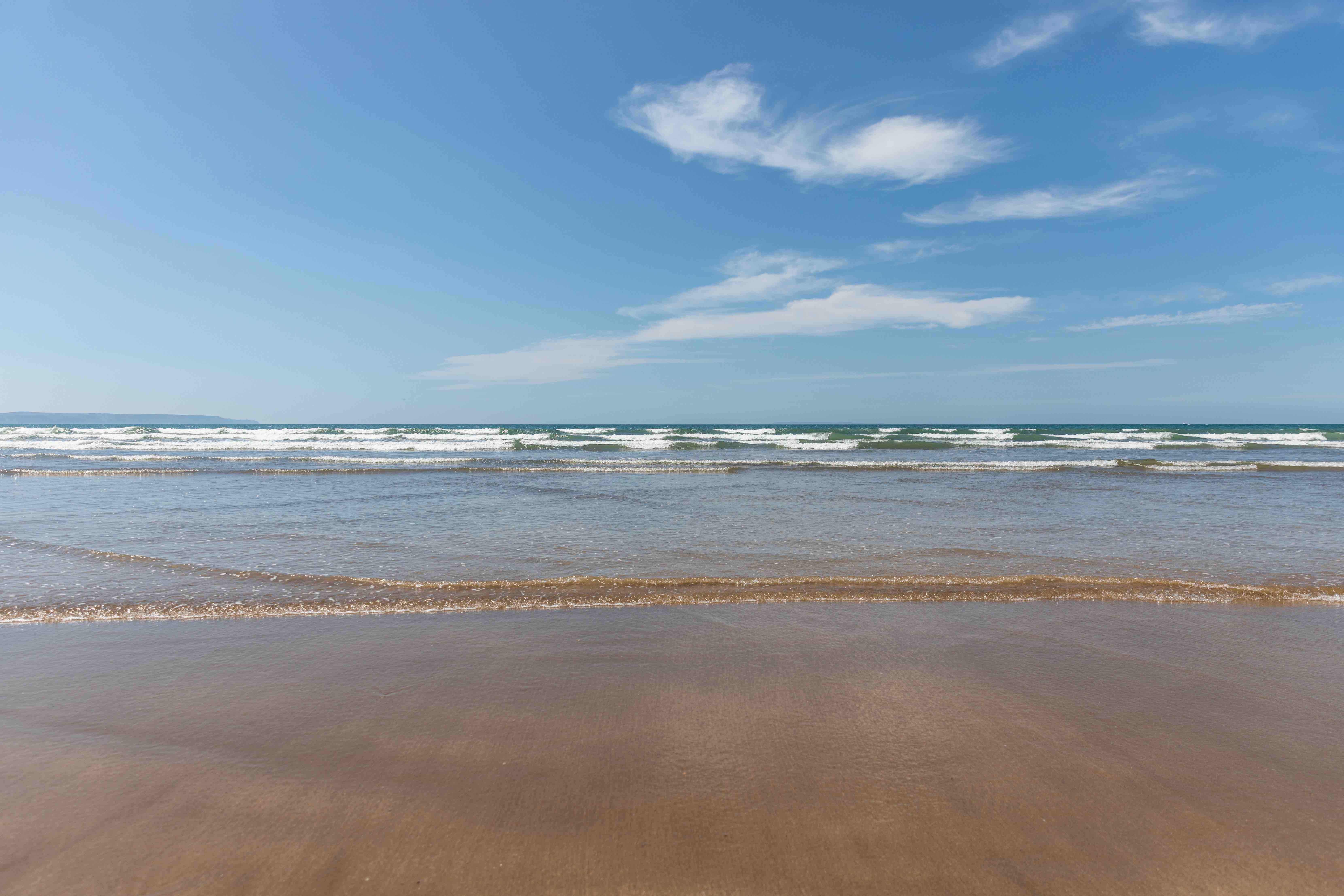 Calm ocean waves gently washing onto a sandy beach under a bright blue sky with scattered clouds.
