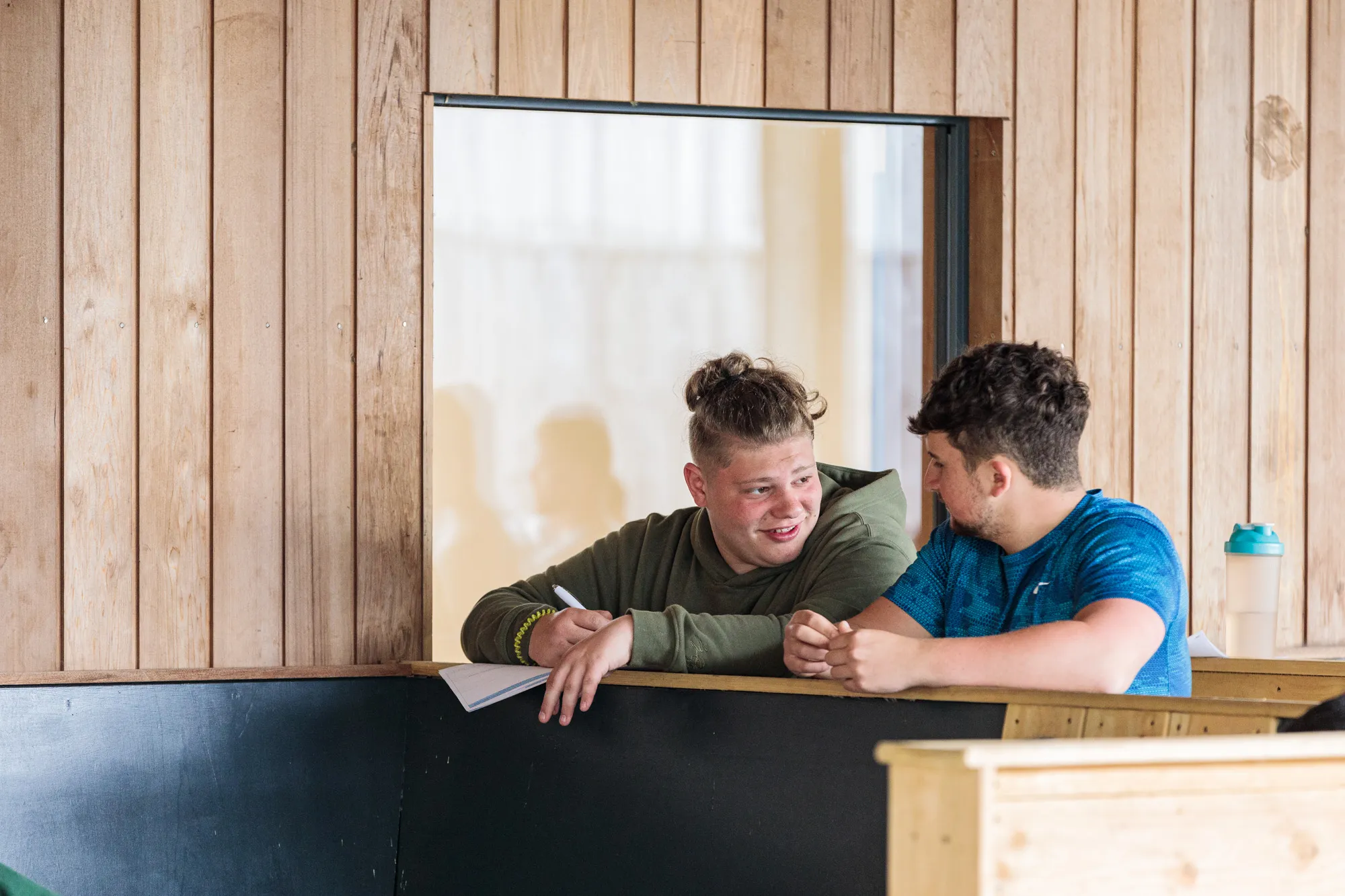 Two young men sitting and talking across a wooden desk, one holding a pen and notebook.