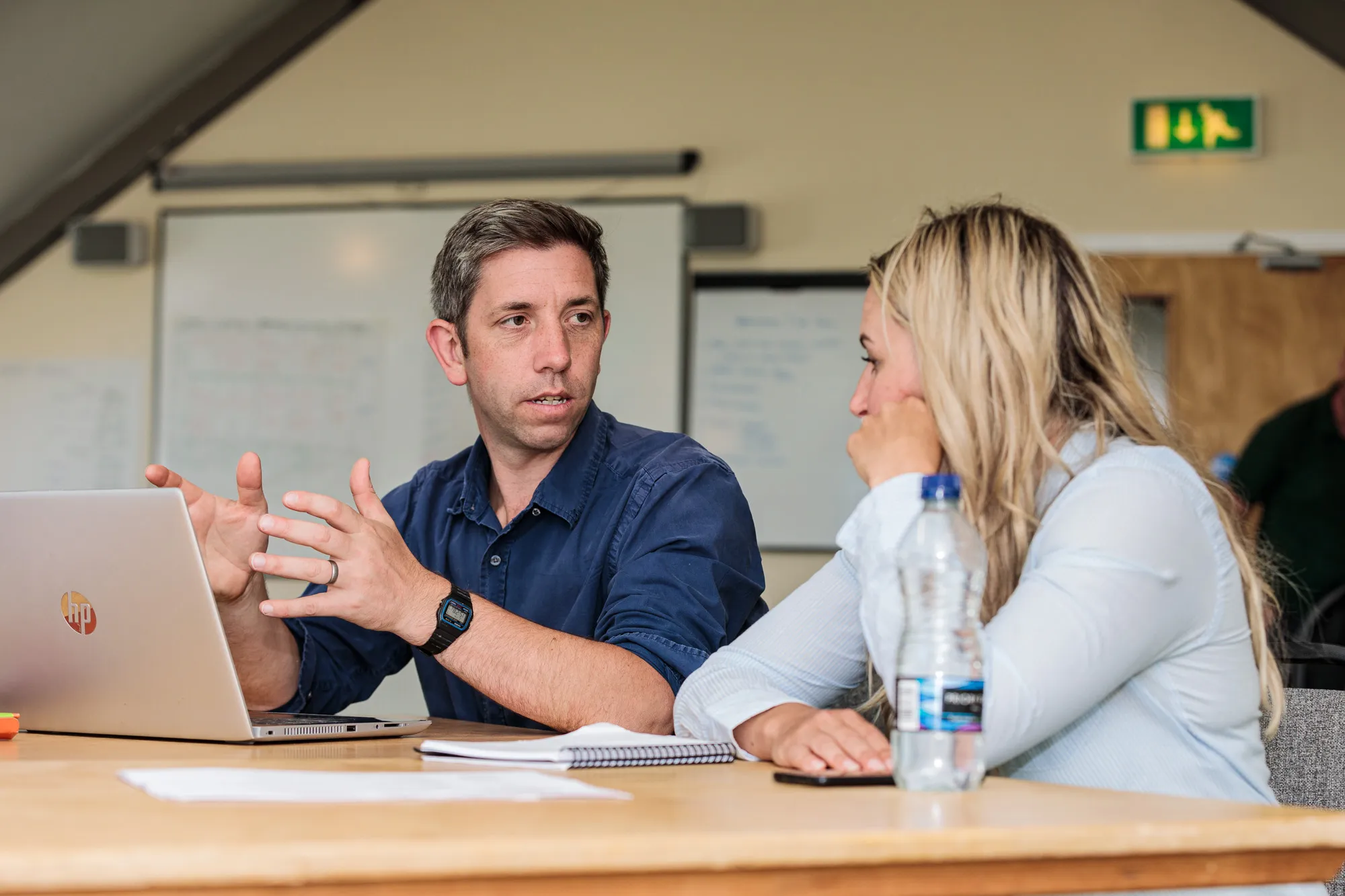 Man explaining something to woman at a table with laptop, notebook, and water bottle in a meeting room.
