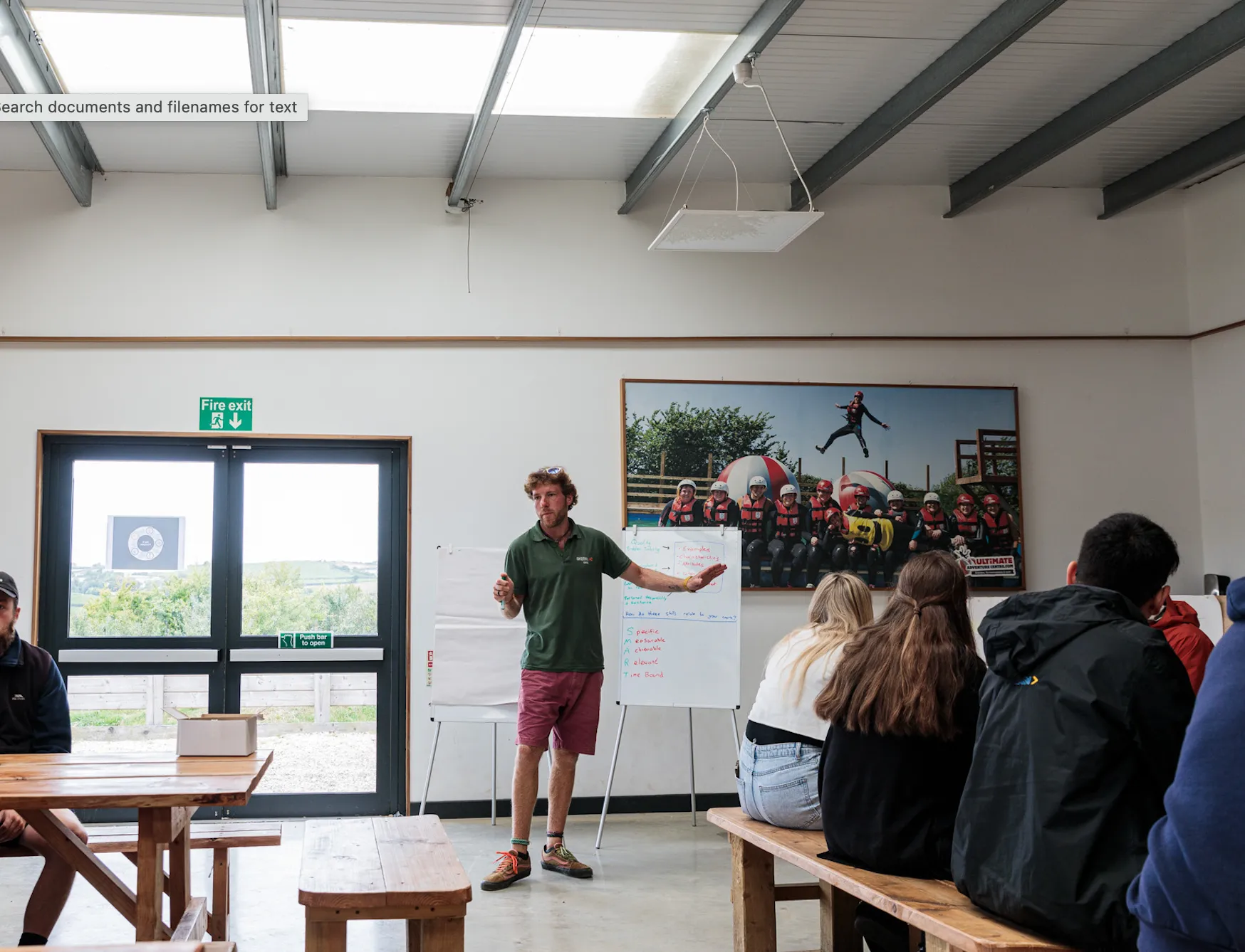 Man giving a presentation in a room with benches, a large photo of people in helmets on a wall, and a fire exit door.