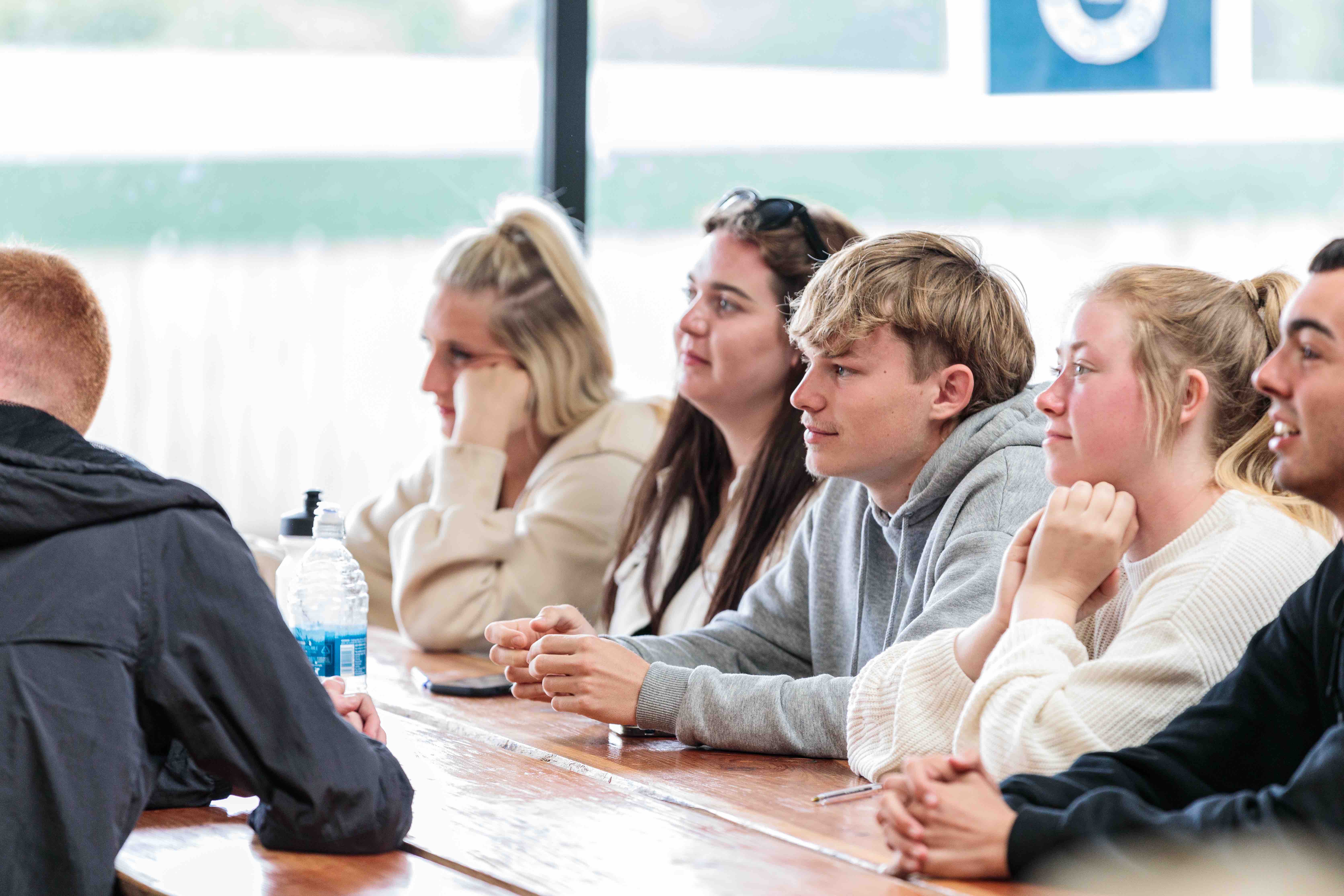Group of young adults attentively listening while seated at a wooden table indoors.