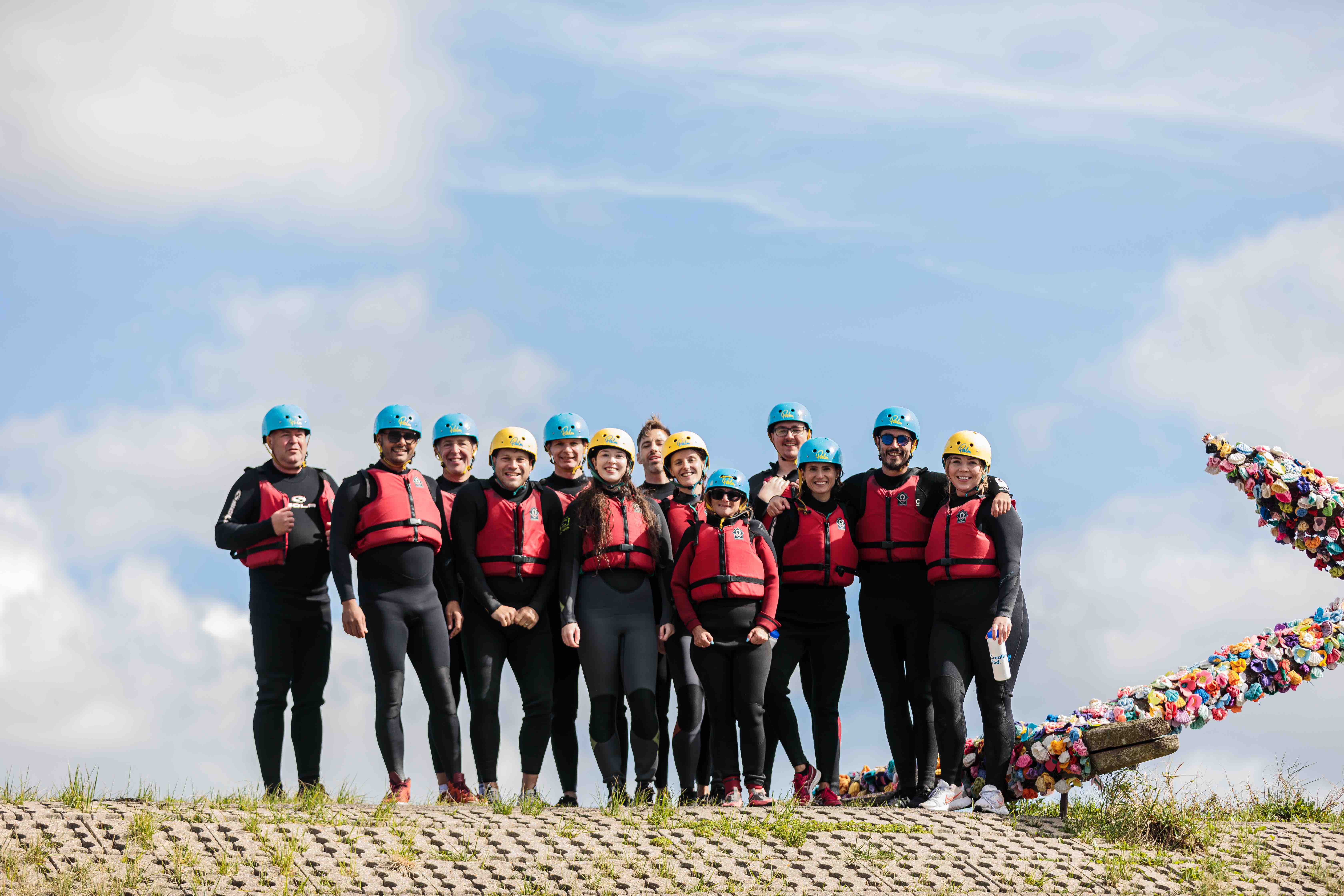 Group of twelve people in wetsuits, red life jackets, and blue or yellow helmets standing outdoors on a paved surface with a cloudy sky background.