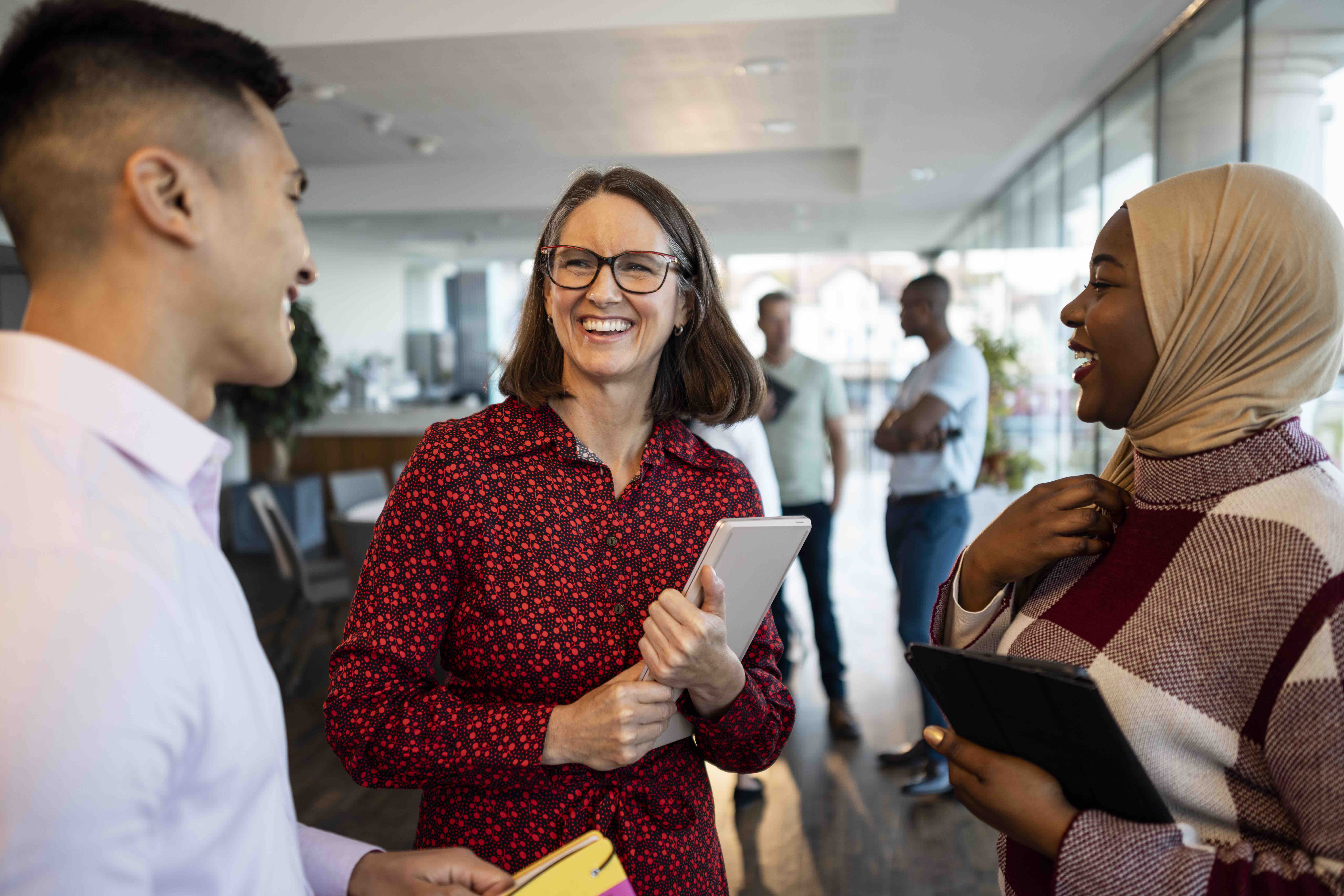 Three diverse colleagues smiling and holding tablets while conversing in a bright office space.