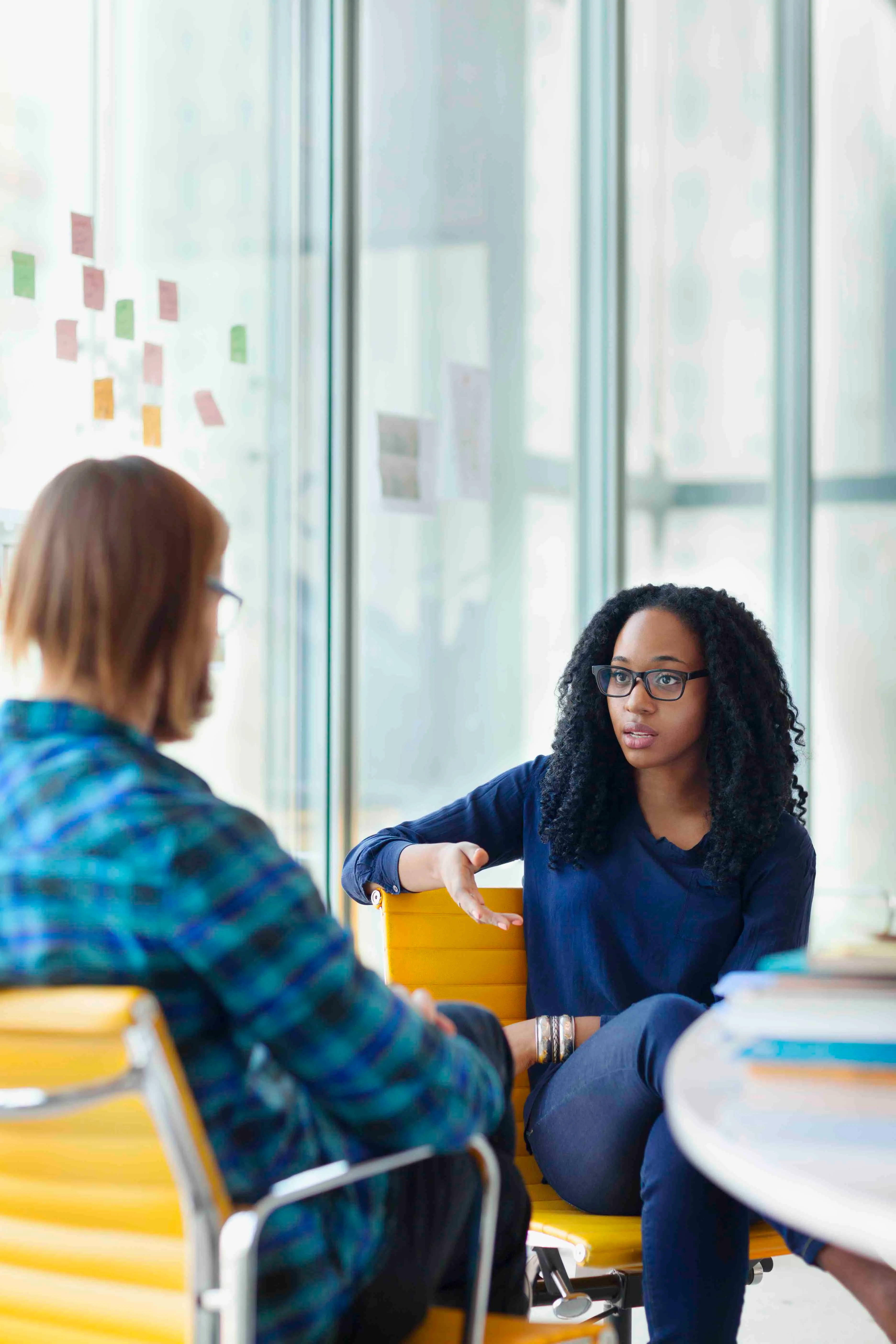 Two women sitting in yellow office chairs having a discussion in a modern glass-walled office.