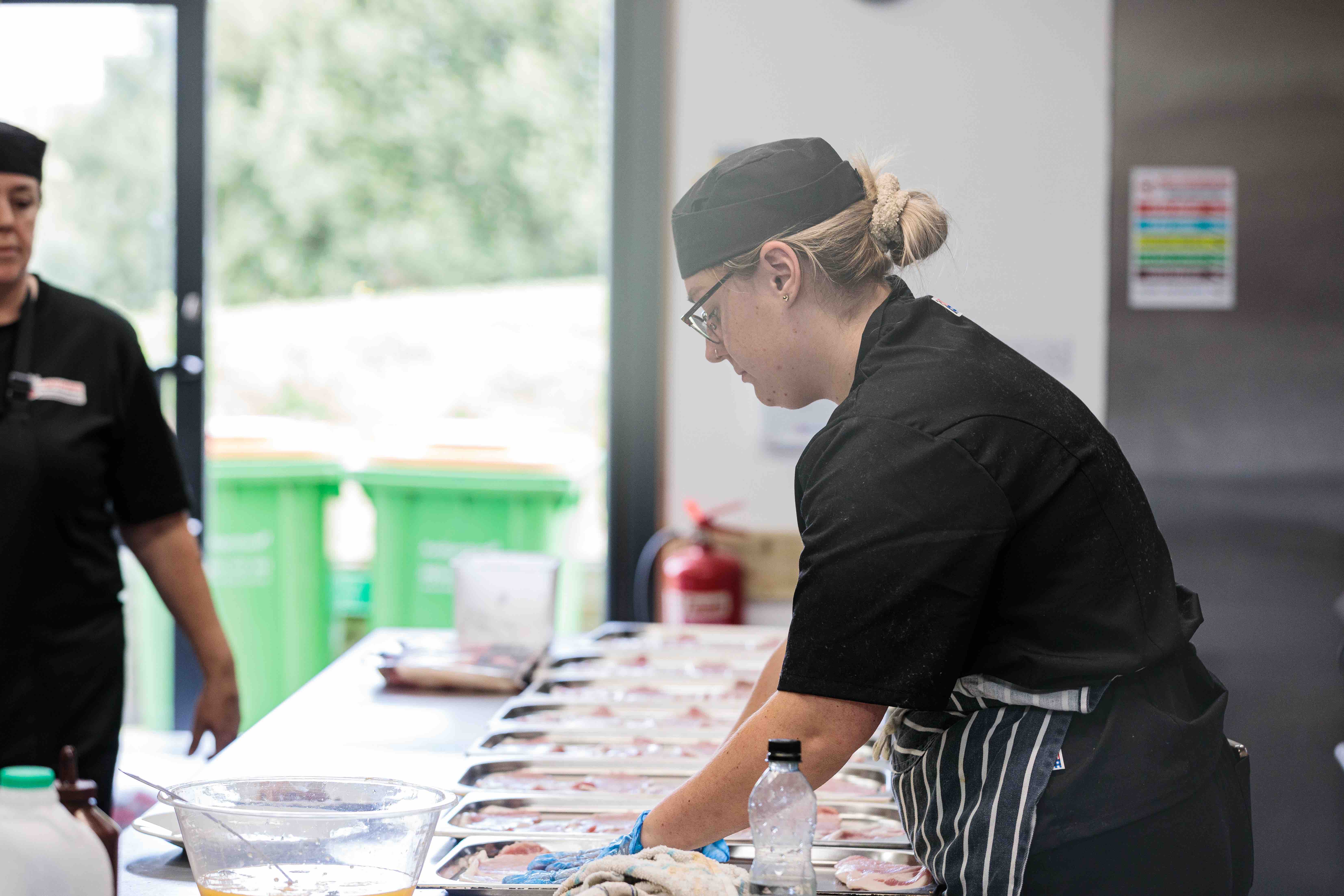 A chef in a black uniform and hat preparing multiple trays of raw meat in a kitchen.