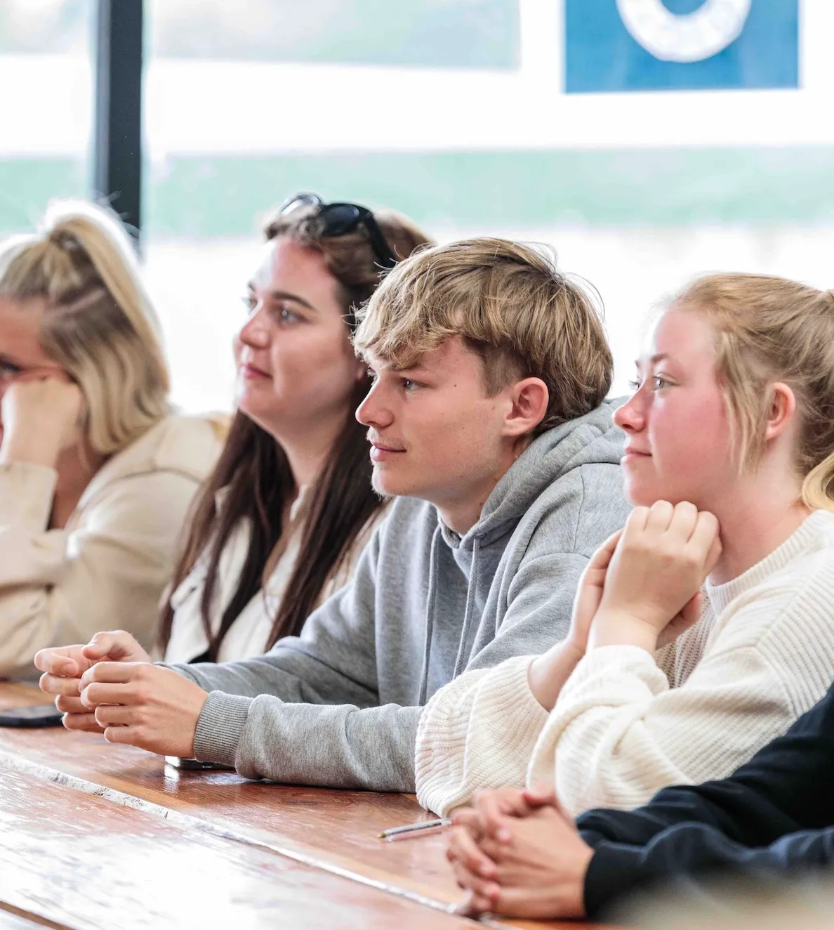 Group of young adults attentively listening while seated at a wooden table in a bright room.