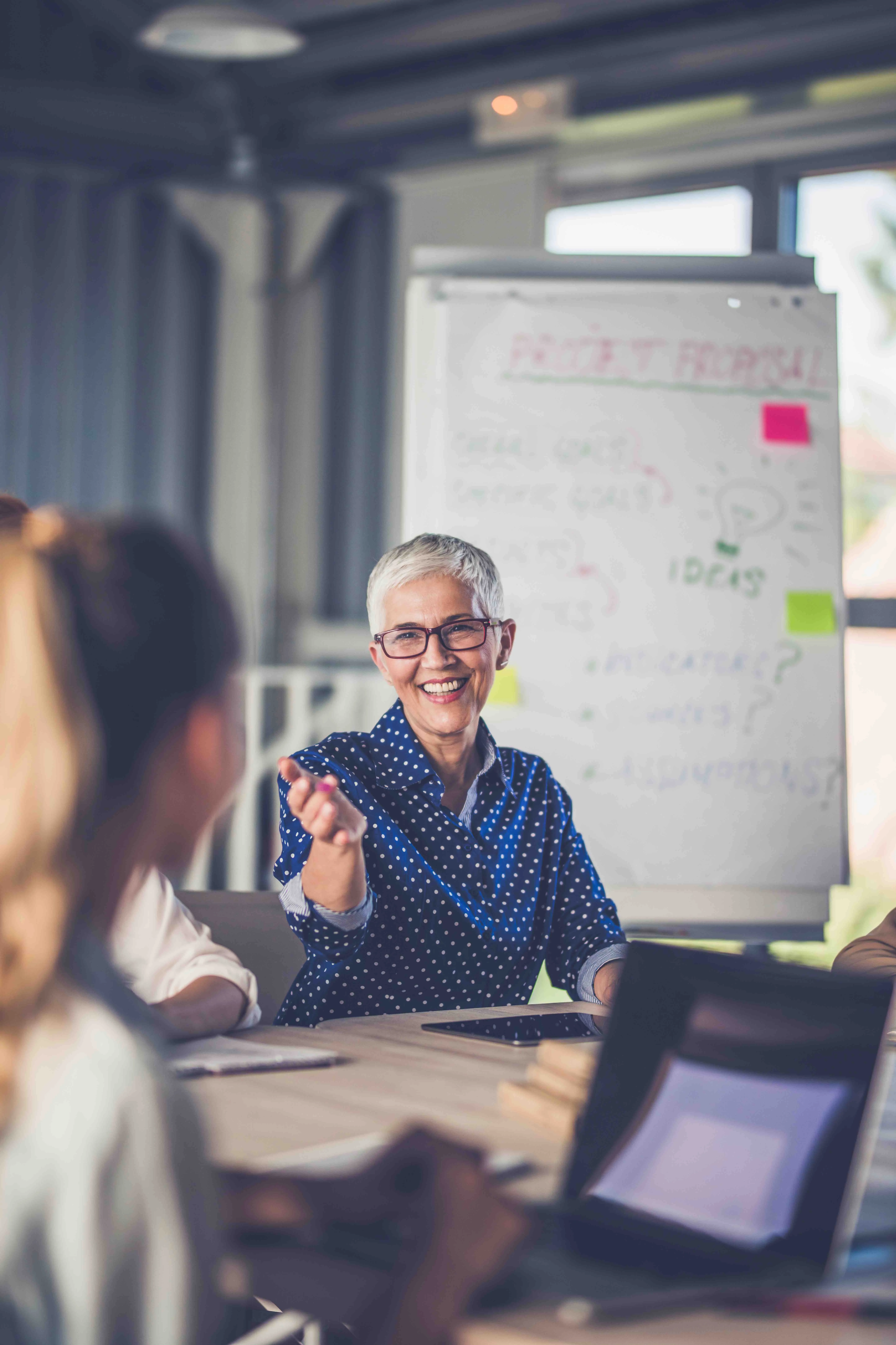 Smiling older woman with short grey hair and glasses engaging with colleagues in a meeting room with a project proposal board in the background.