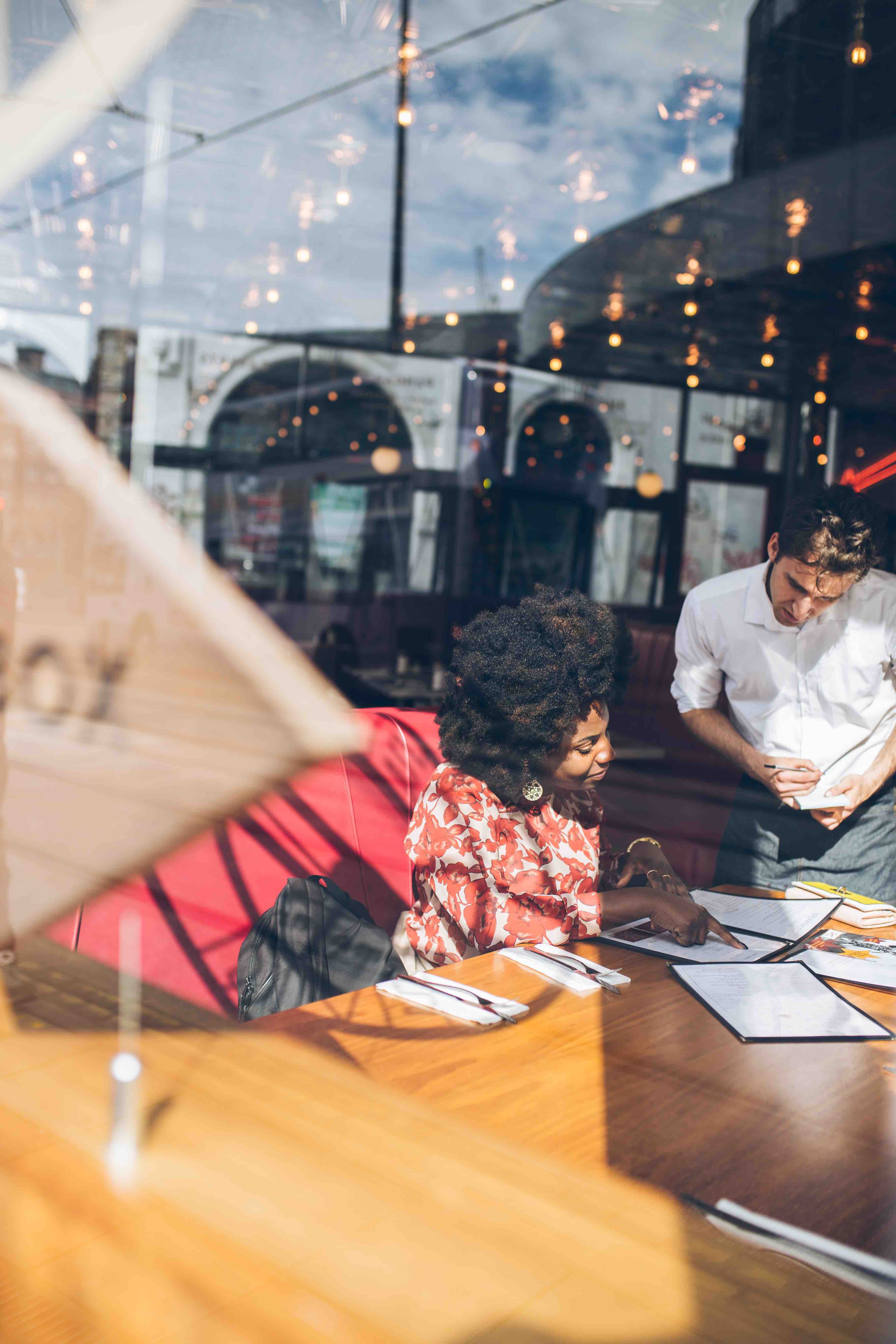 A woman in a floral blouse points to a menu while a waiter in a white shirt takes her order at a cafe with glass windows and string lights.