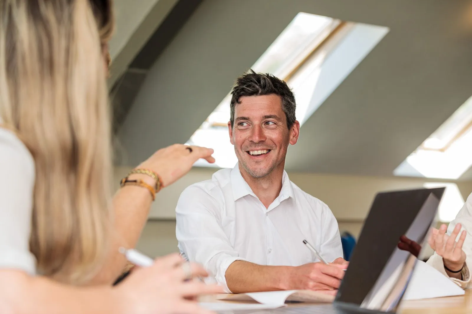 Smiling man in white shirt listening and engaging in conversation during a meeting at a table with a laptop and documents.