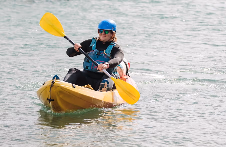 Person wearing a blue helmet and life vest paddling a yellow kayak on calm water.