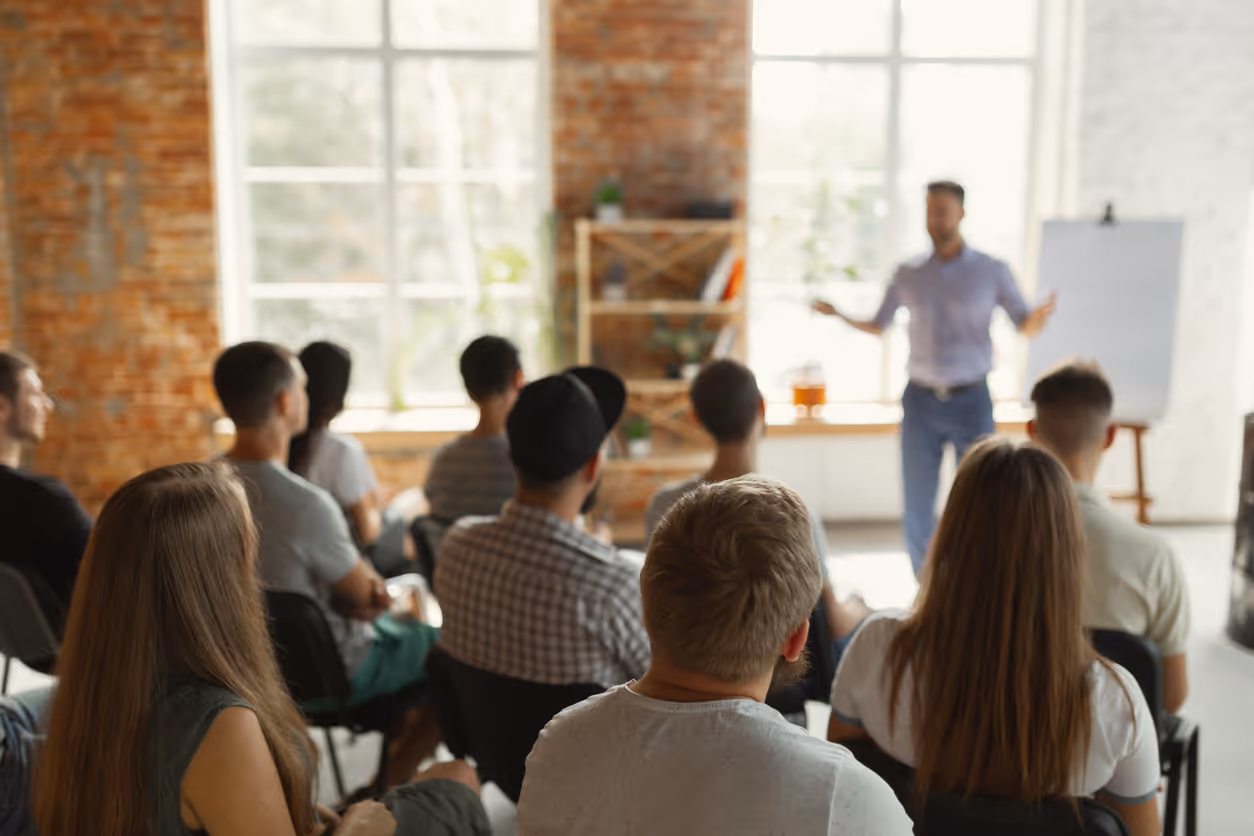 Group of people seated facing a man giving a presentation in a bright room with large windows and a flip chart.