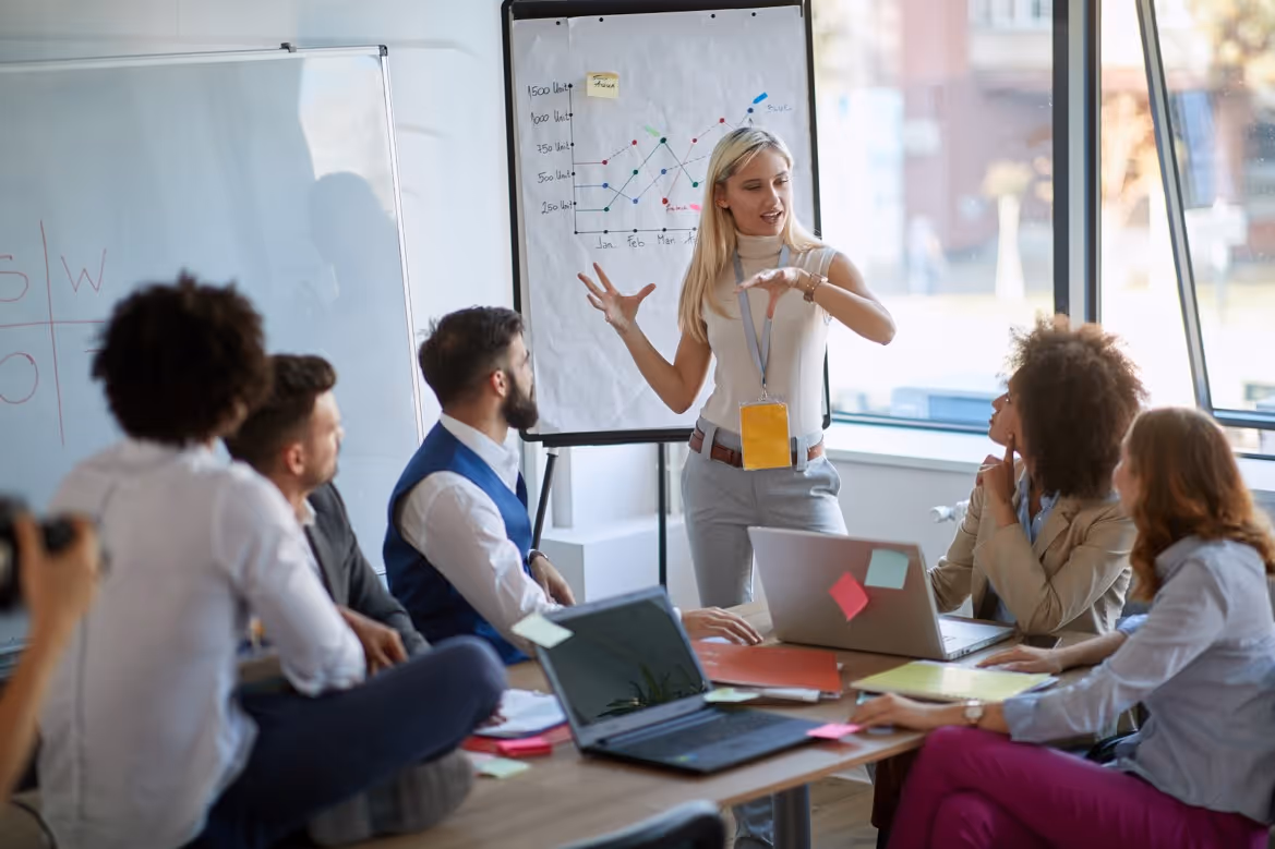 A blonde woman presents business data on a flipchart to a diverse group seated around a table with laptops and notes.