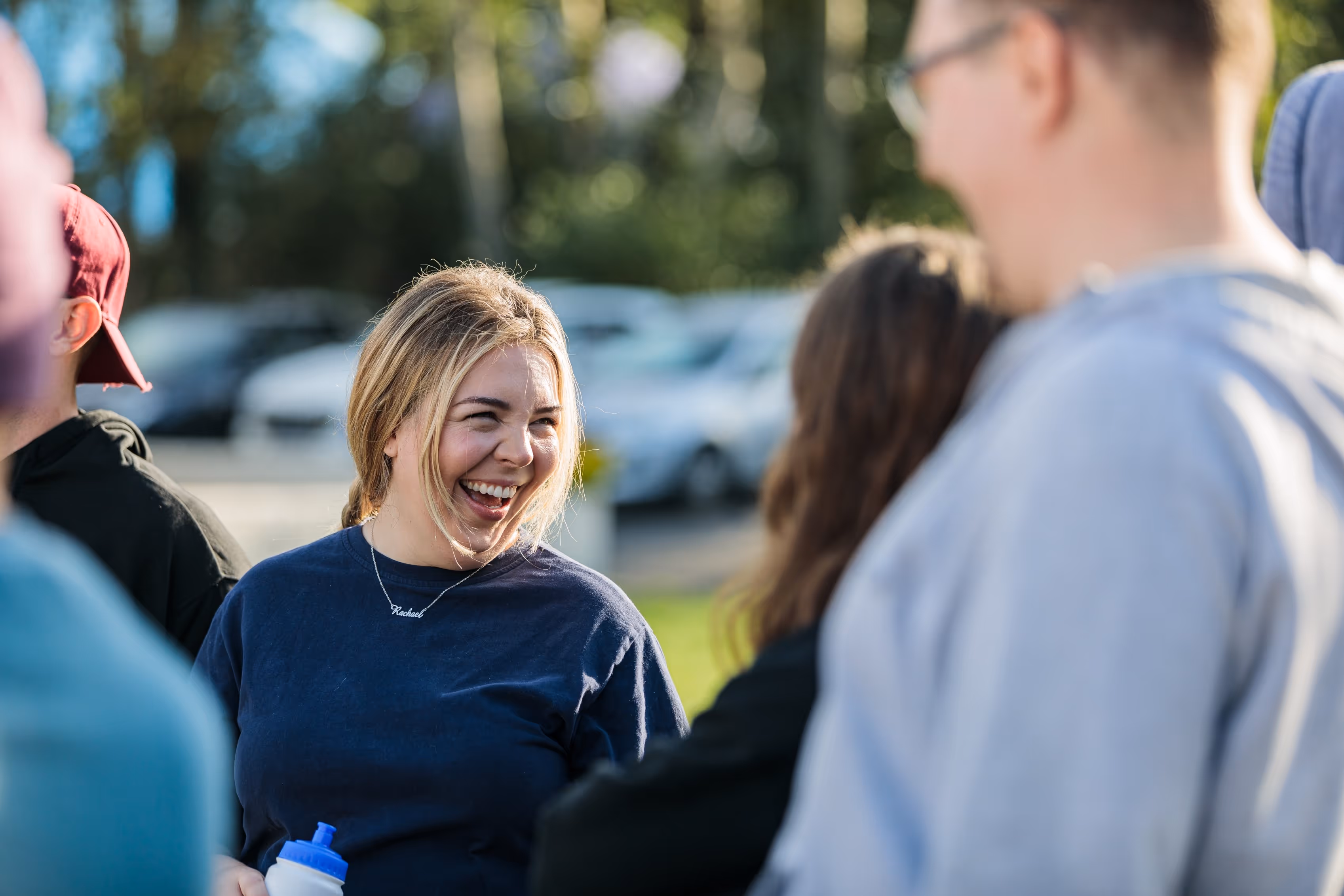 A young woman with blonde hair smiling and holding a water bottle while talking with others outdoors.