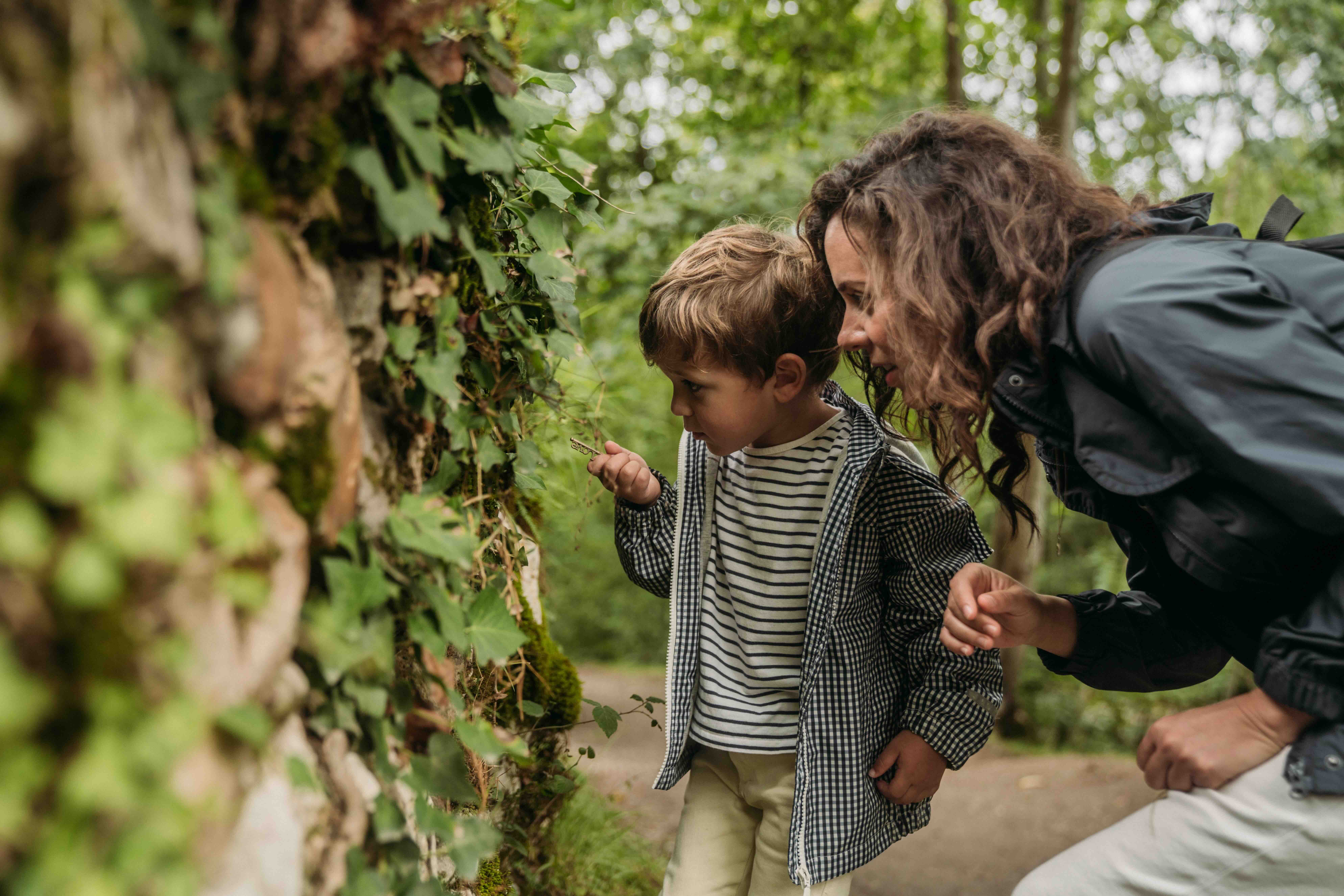 Child examining leaves on a mossy wall outdoors with an adult woman crouching beside him.