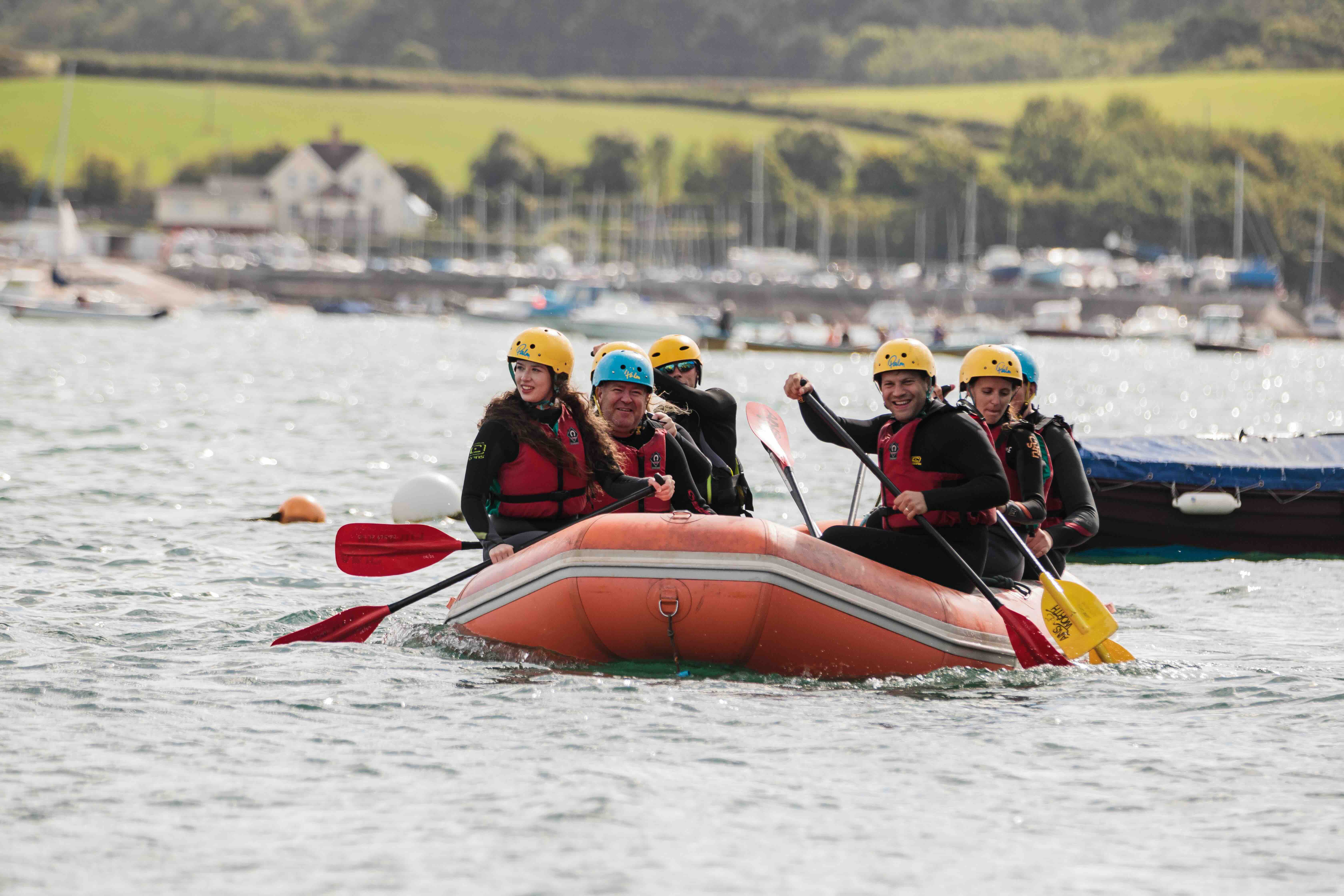 Six people wearing helmets and life jackets paddling an orange inflatable raft on a body of water near a marina.