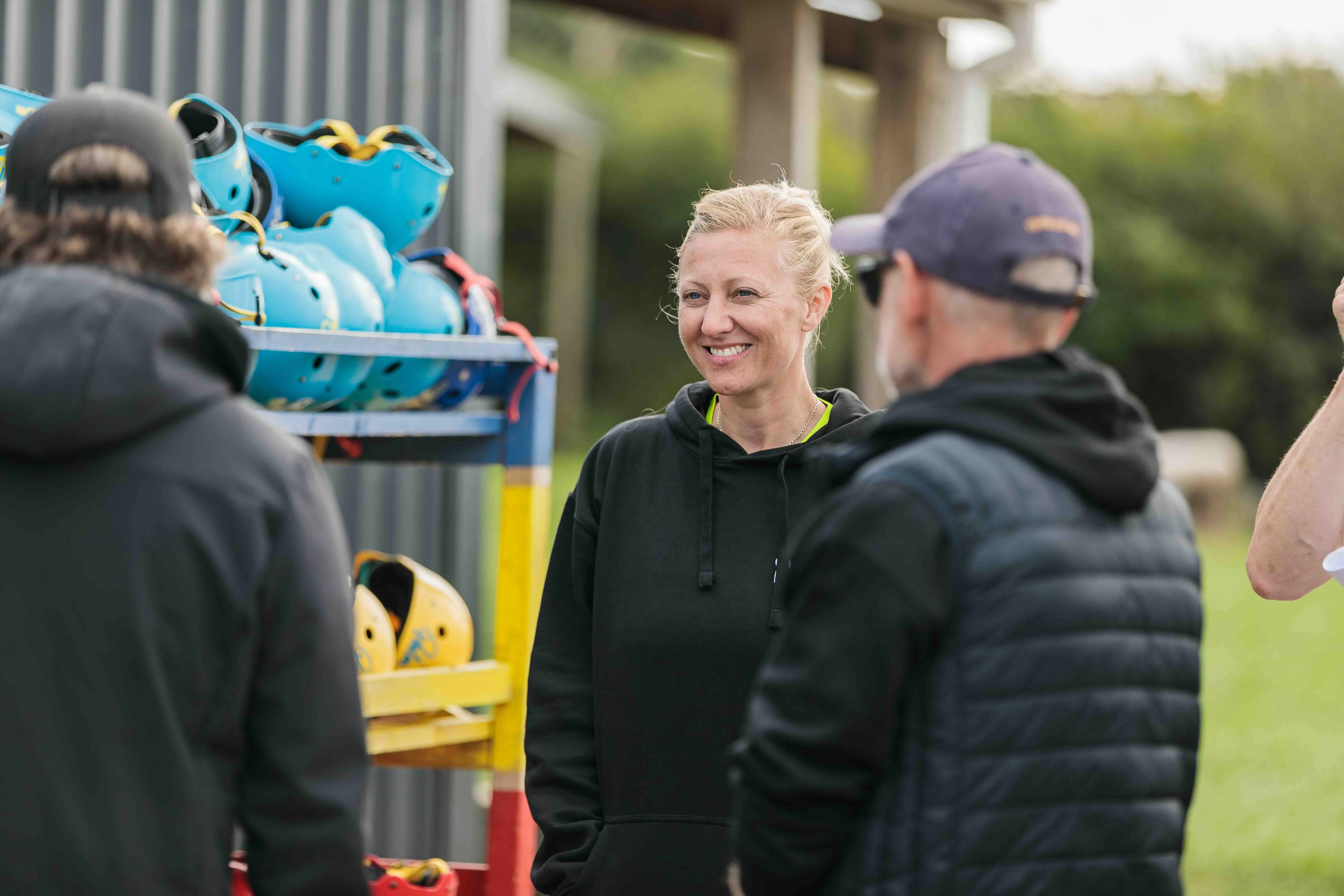 A smiling blonde woman in a black hoodie stands outdoors next to a rack of blue and yellow helmets, talking with two men.
