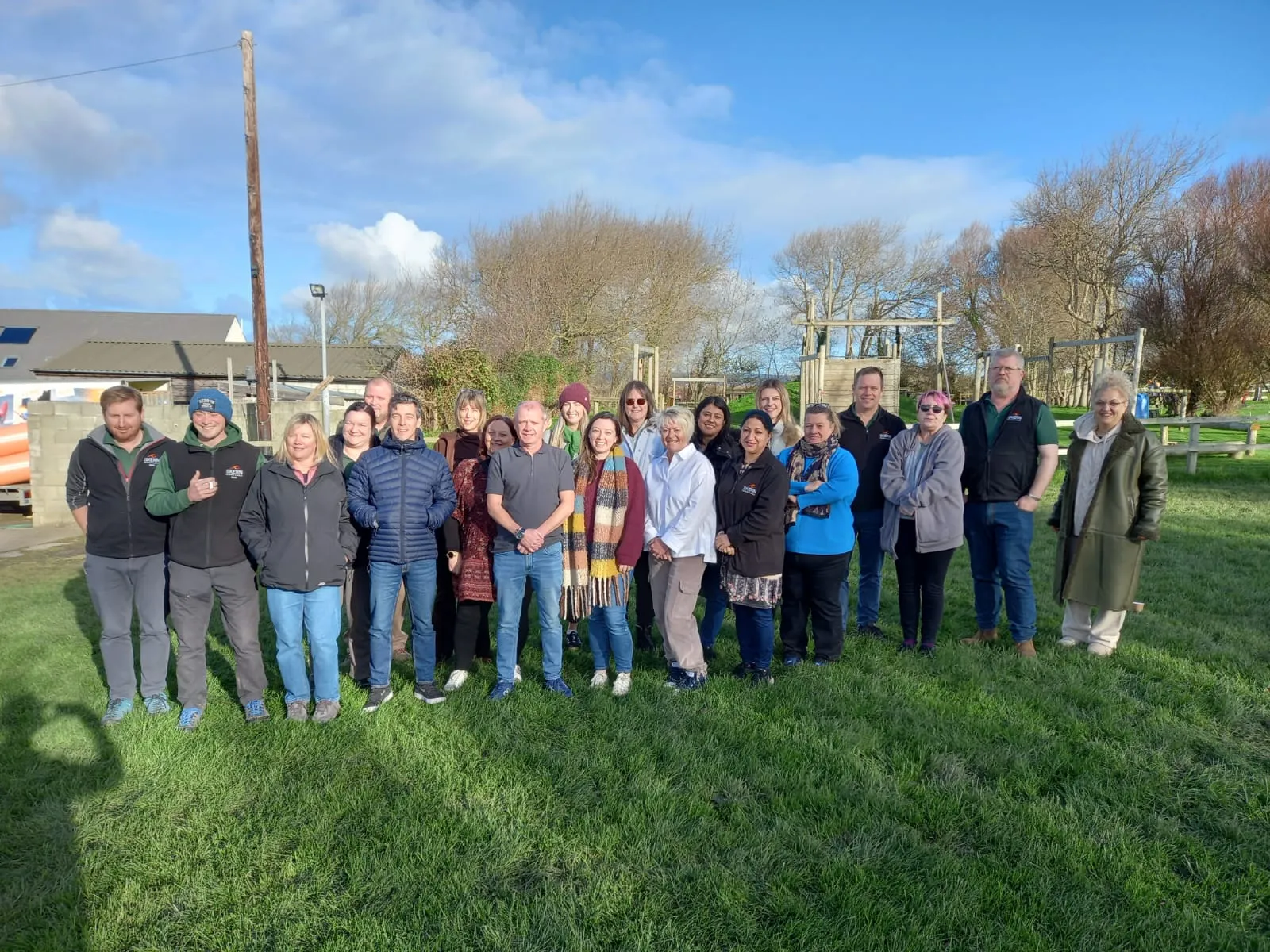 Group of 20 people standing on grass outdoors, posing for a photo under a blue sky with some clouds.