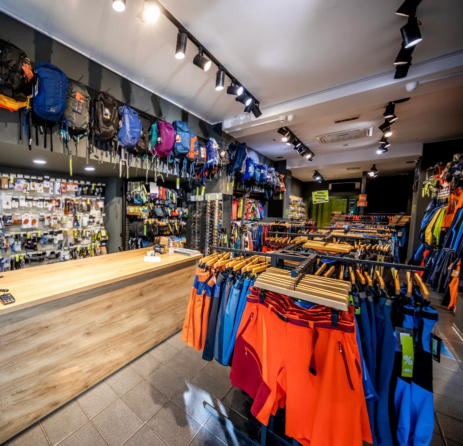 Outdoor retail store interior with backpacks hanging on the wall, racks of colorful hiking and outdoor clothing, and a wooden checkout counter.