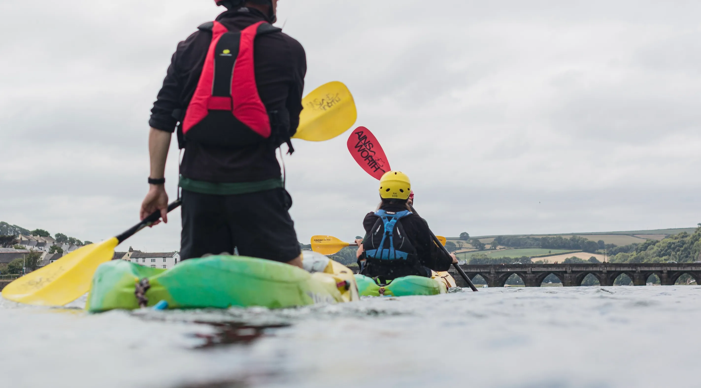 Two people kayaking on a river with green hills and a stone bridge in the background.