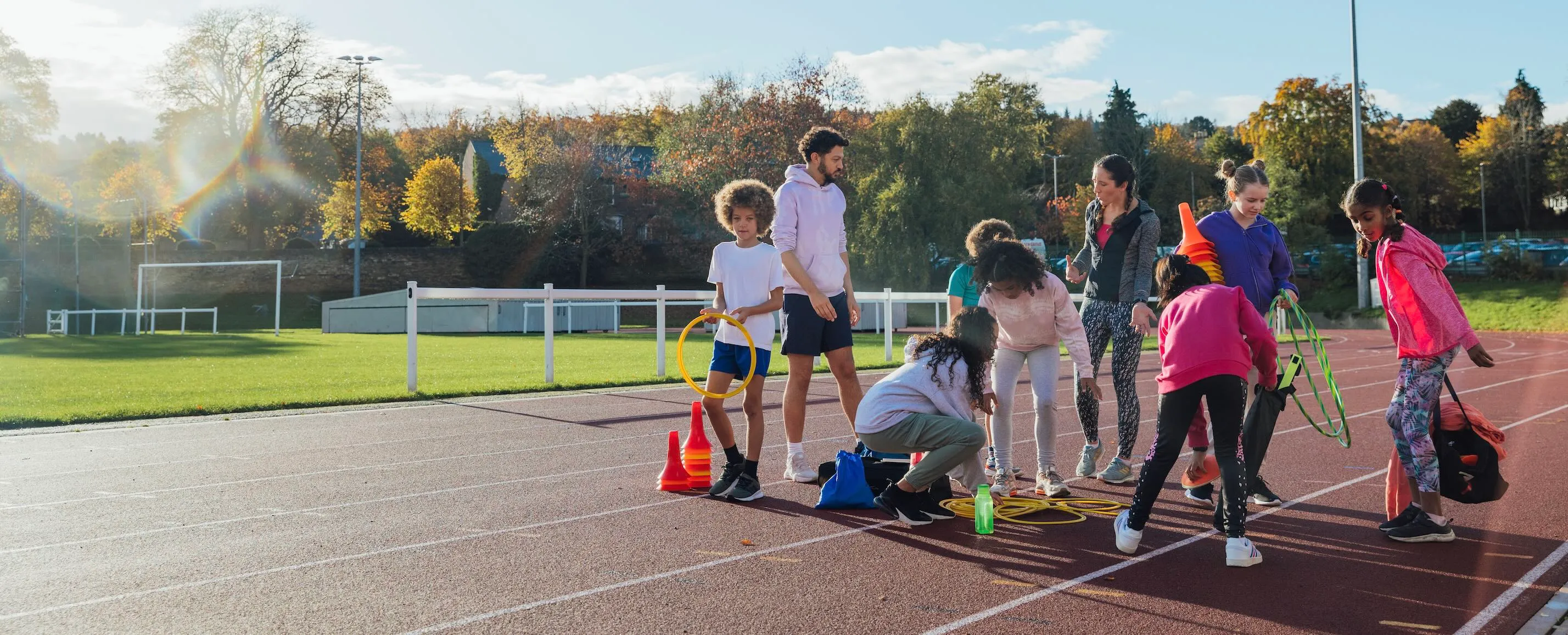 A group of children and two coaches organizing sports equipment on an outdoor running track in autumn.