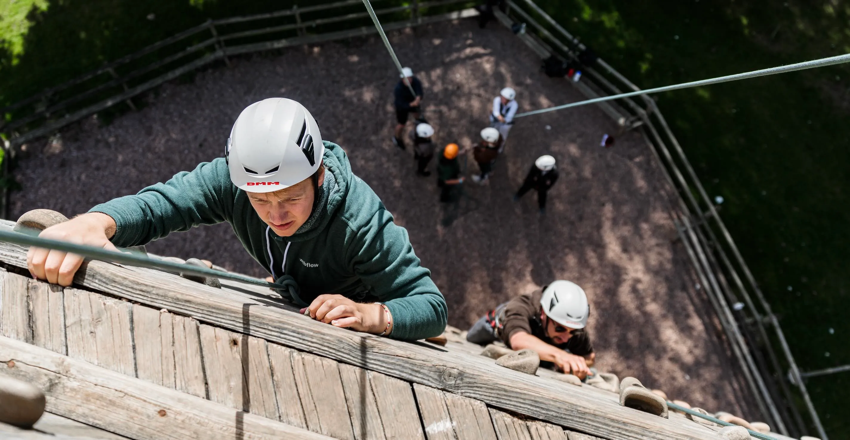 Two climbers wearing helmets and harnesses ascending a wooden climbing wall outdoors with a group of people below.