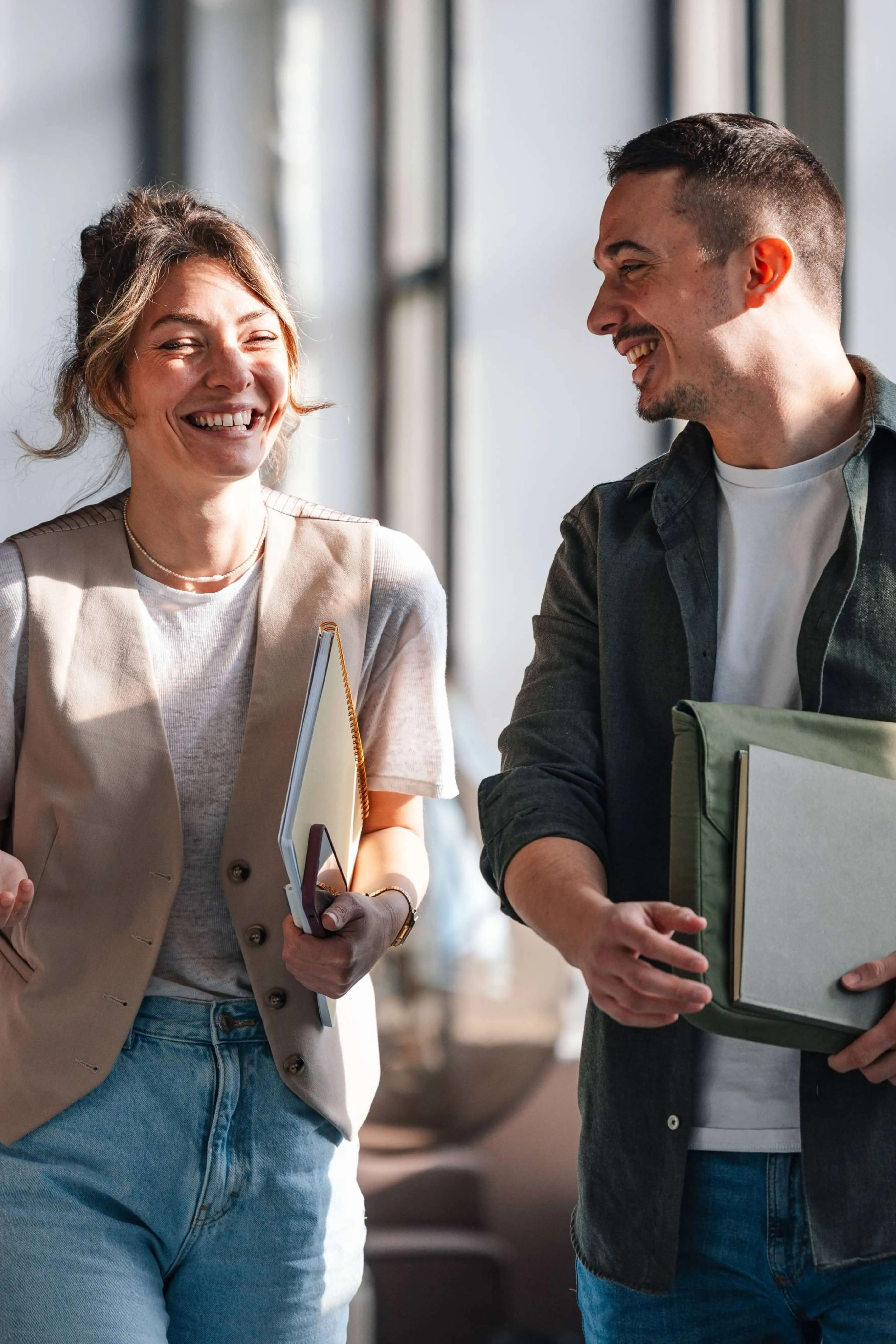 A smiling woman and man walking indoors, each holding notebooks and folders while engaging in conversation.