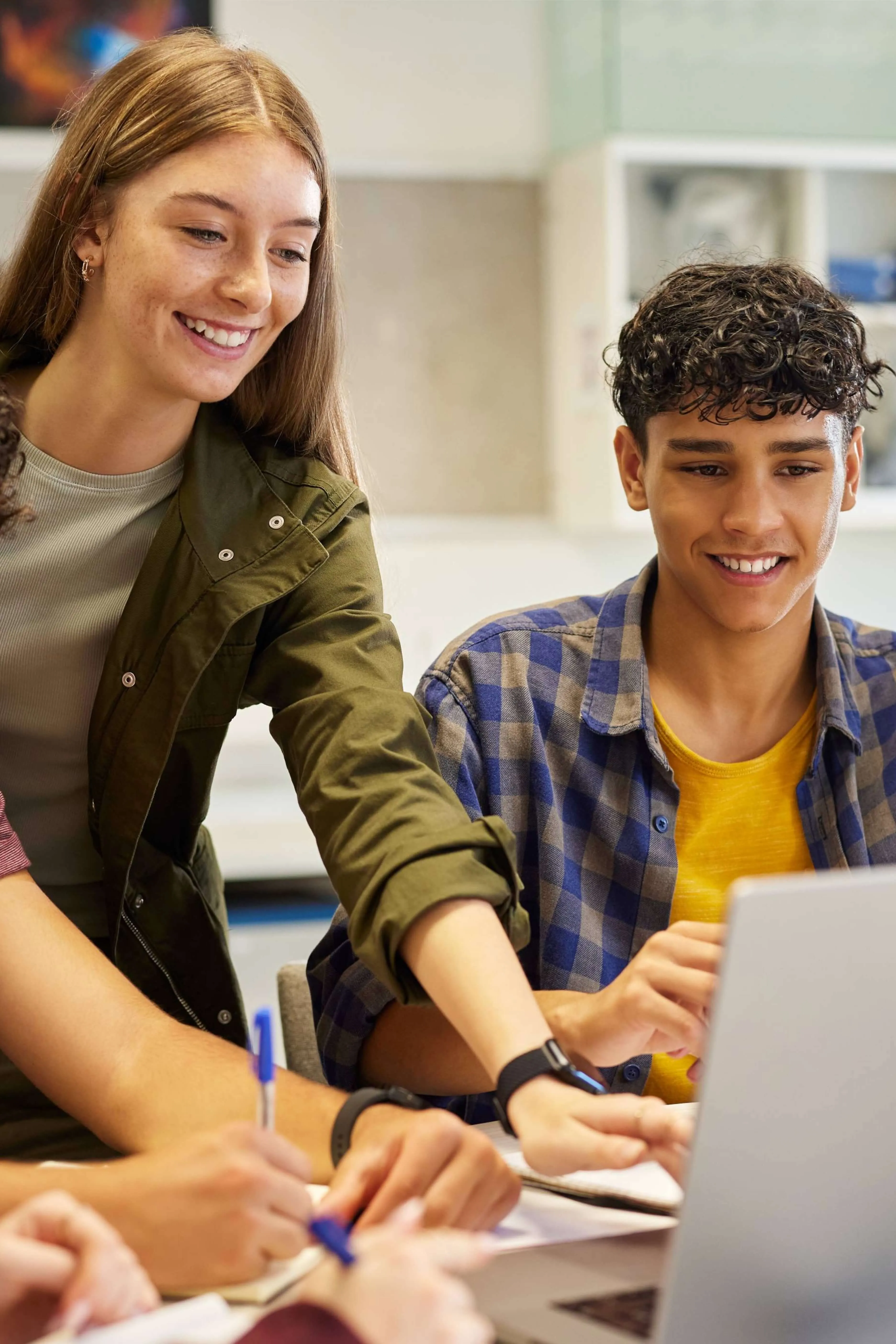 Two smiling teenagers, one pointing at a laptop screen while the other looks on, engaged in a group study session.