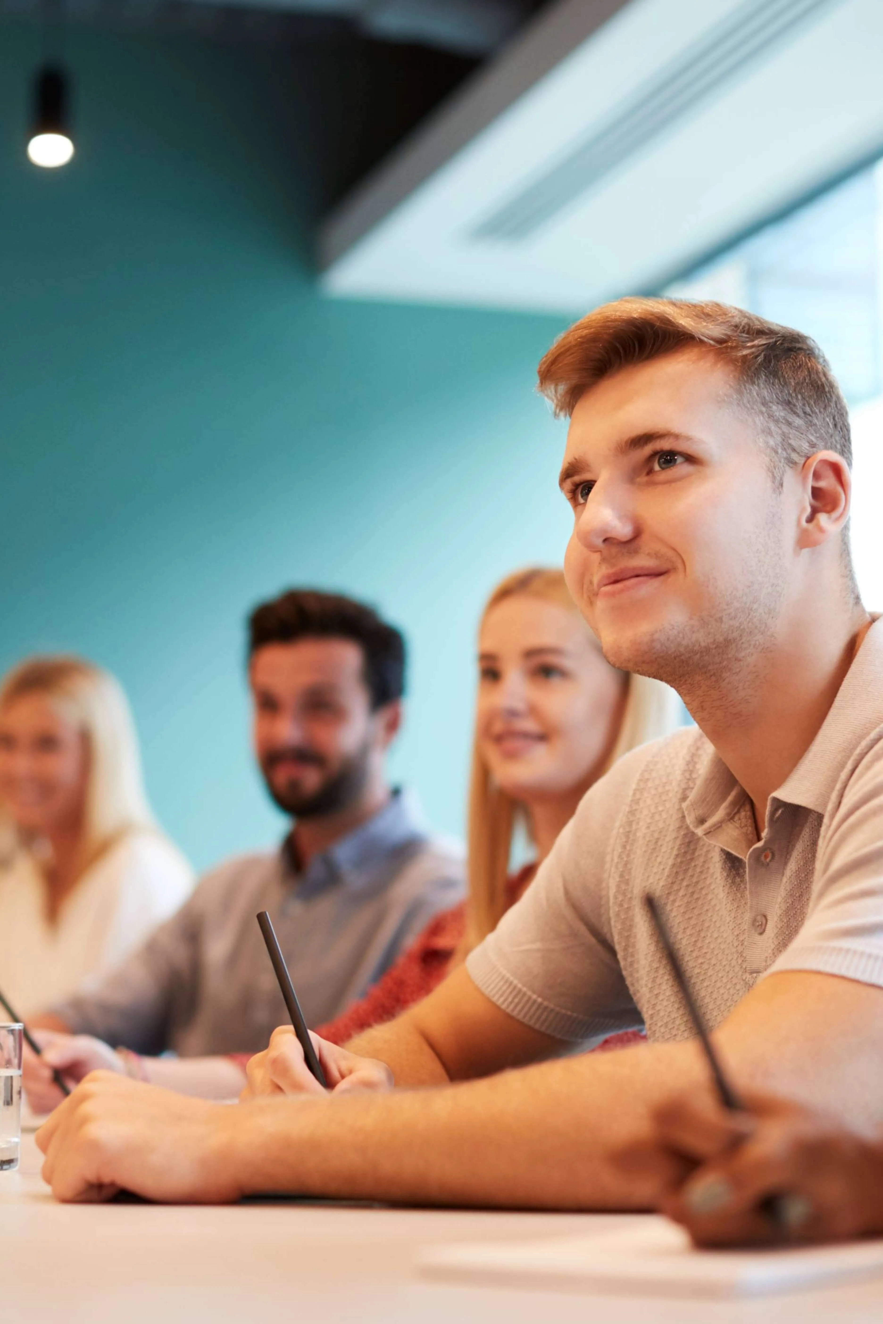 A group of young adults sitting at a table attentively listening and taking notes in a classroom or meeting setting.