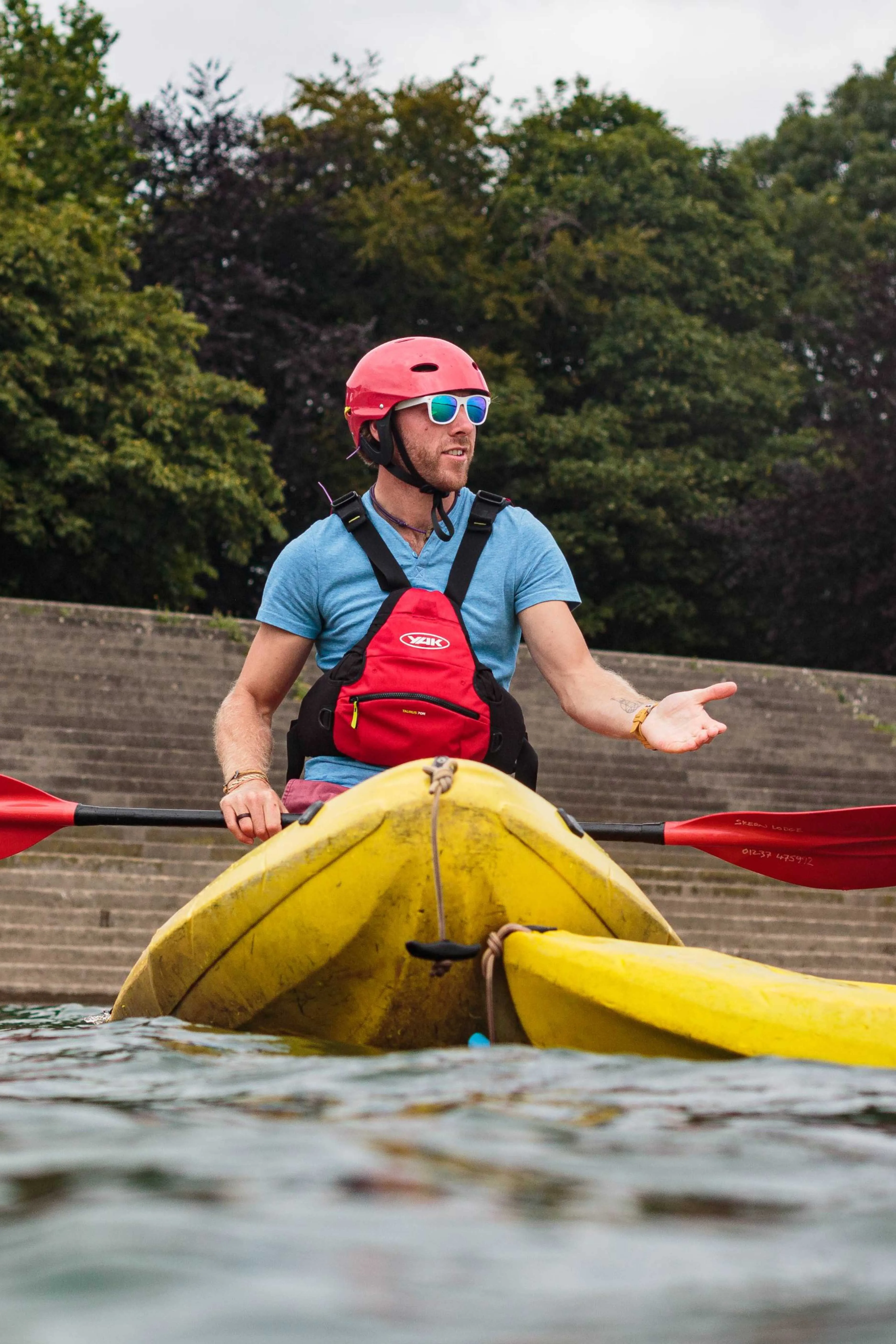 Man wearing a red helmet and blue shirt paddling a yellow kayak on water with trees and concrete steps in the background.