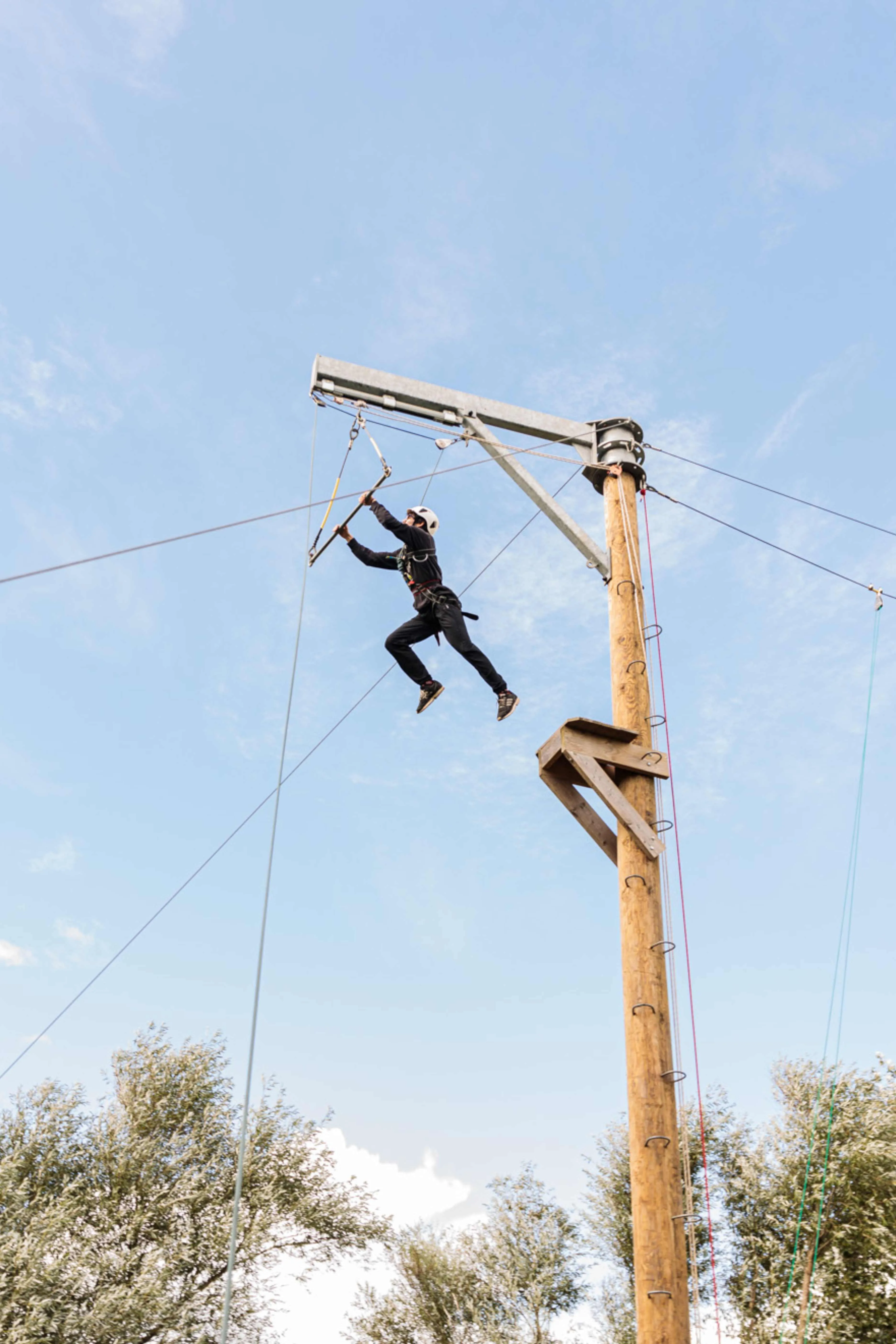Person wearing safety gear jumping mid-air from a wooden pole platform in an outdoor ropes course.